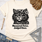 Women's funny woodworking graphic tee shirt for female woodworkers, makers, and DIY builders — Sawdust & Ponytails. Cream t-shirt with a black graphic of a cat wearing goggles and a flower, text "Measured Twice. Judged Once.