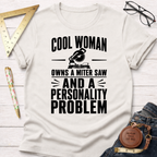 White cotton t-shirt with "Cool Woman Owns A Miter Saw And A Personality Problem" graphic.