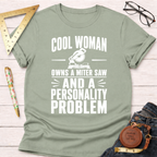 Sage green cotton t-shirt with "Cool Woman Owns A Miter Saw And A Personality Problem" graphic.