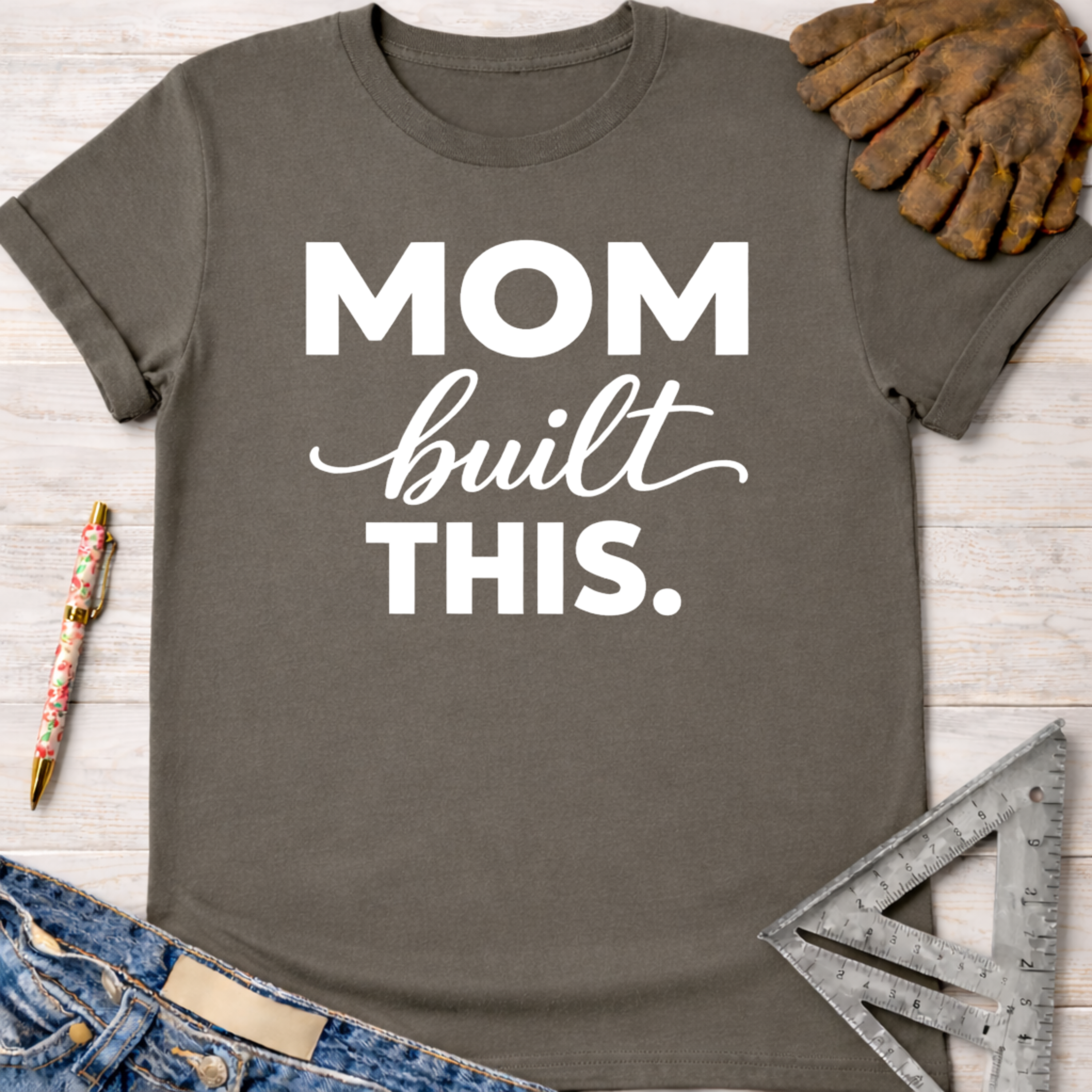 Charcoal gray "Mom built THIS." graphic tee with rolled sleeves, next to work gloves and tools. Women's funny woodworking graphic tee shirt for female woodworkers, makers, and DIY builders — Sawdust & Ponytails.