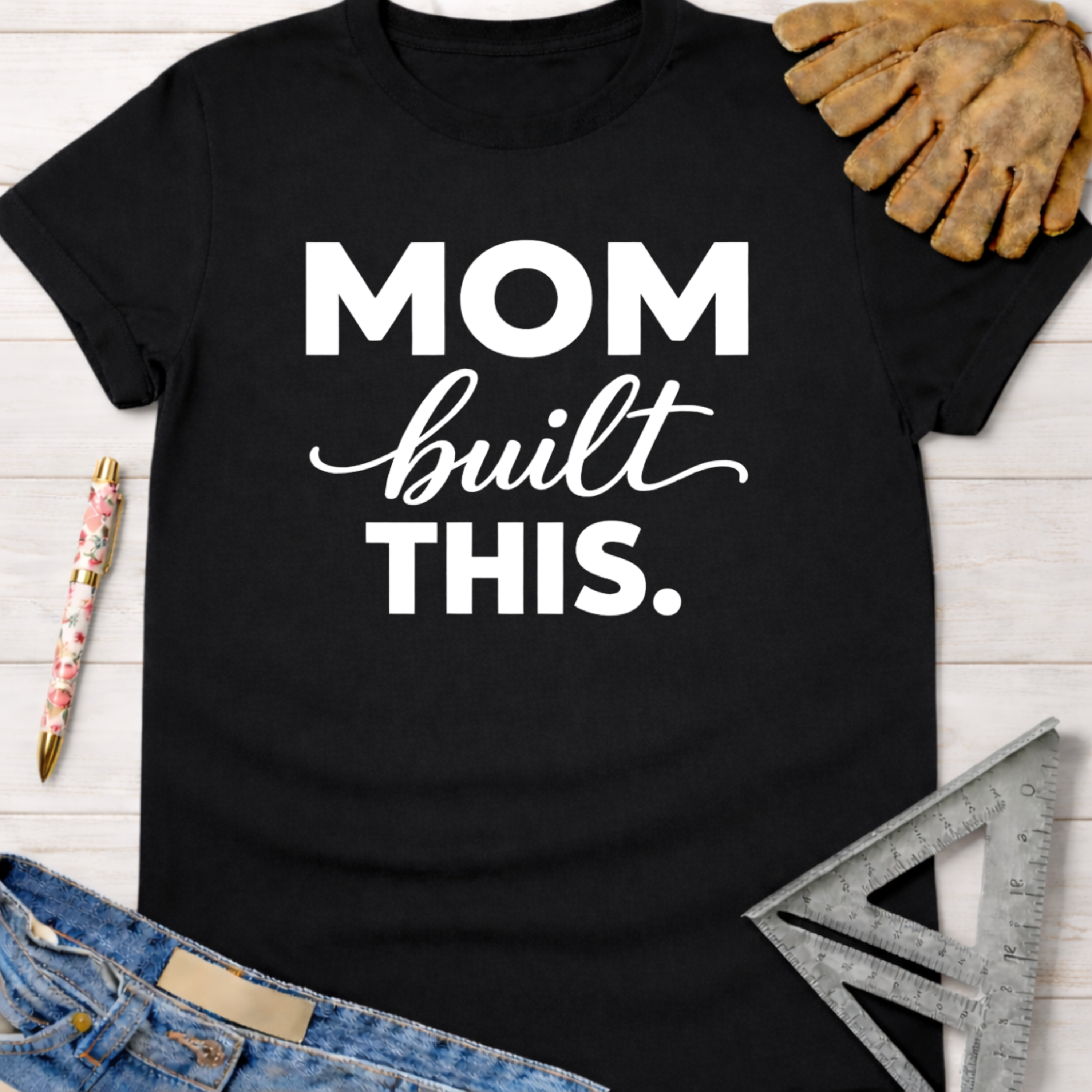 Black cotton t-shirt with "MOM built THIS." in white text, alongside work gloves and a ruler. Women's funny woodworking graphic tee shirt for female woodworkers, makers, and DIY builders — Sawdust & Ponytails