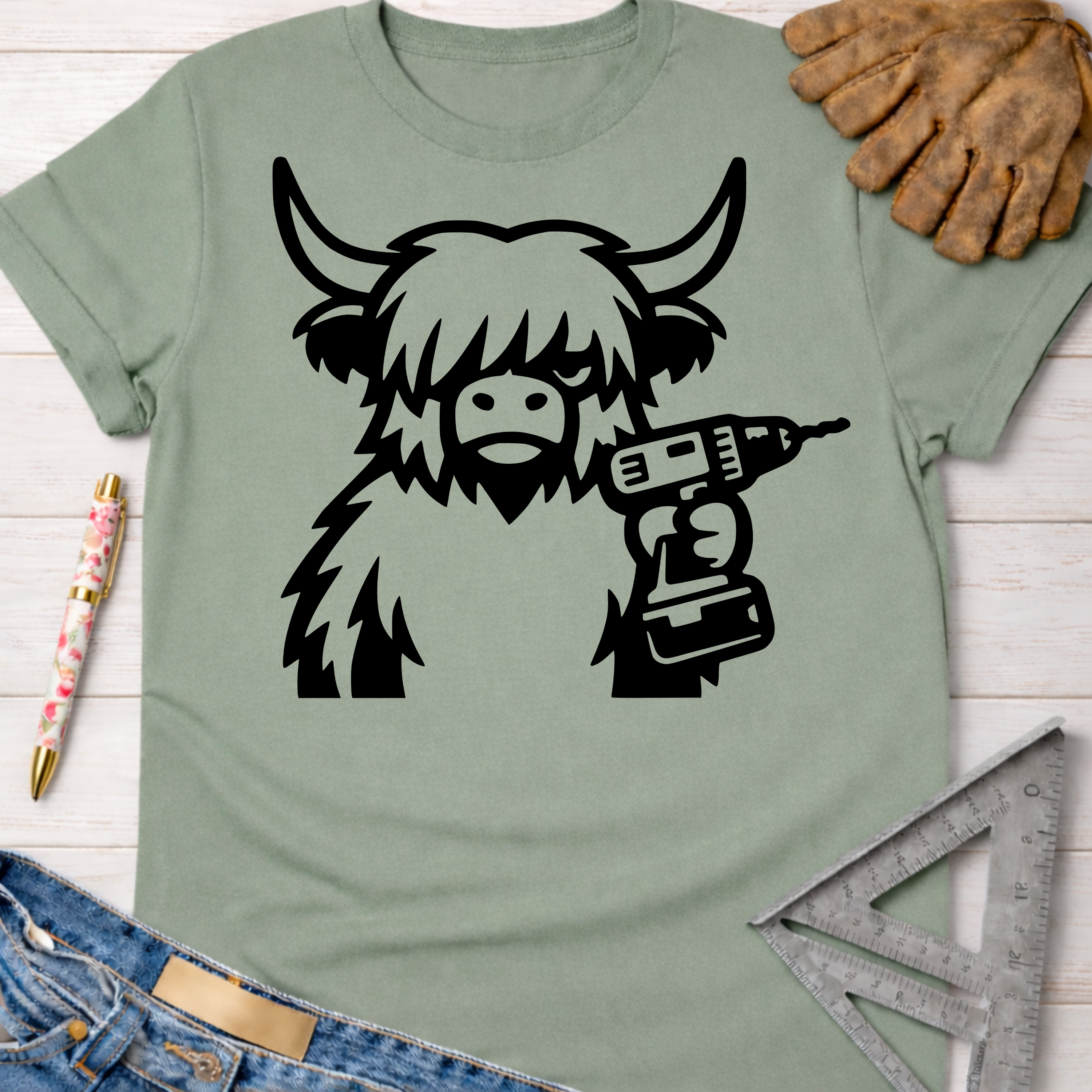 Women's funny woodworking graphic tee shirt for female woodworkers, makers, and DIY builders — Sawdust & Ponytails. Sage green t-shirt with a black graphic of a shaggy Highland cow holding a power drill.