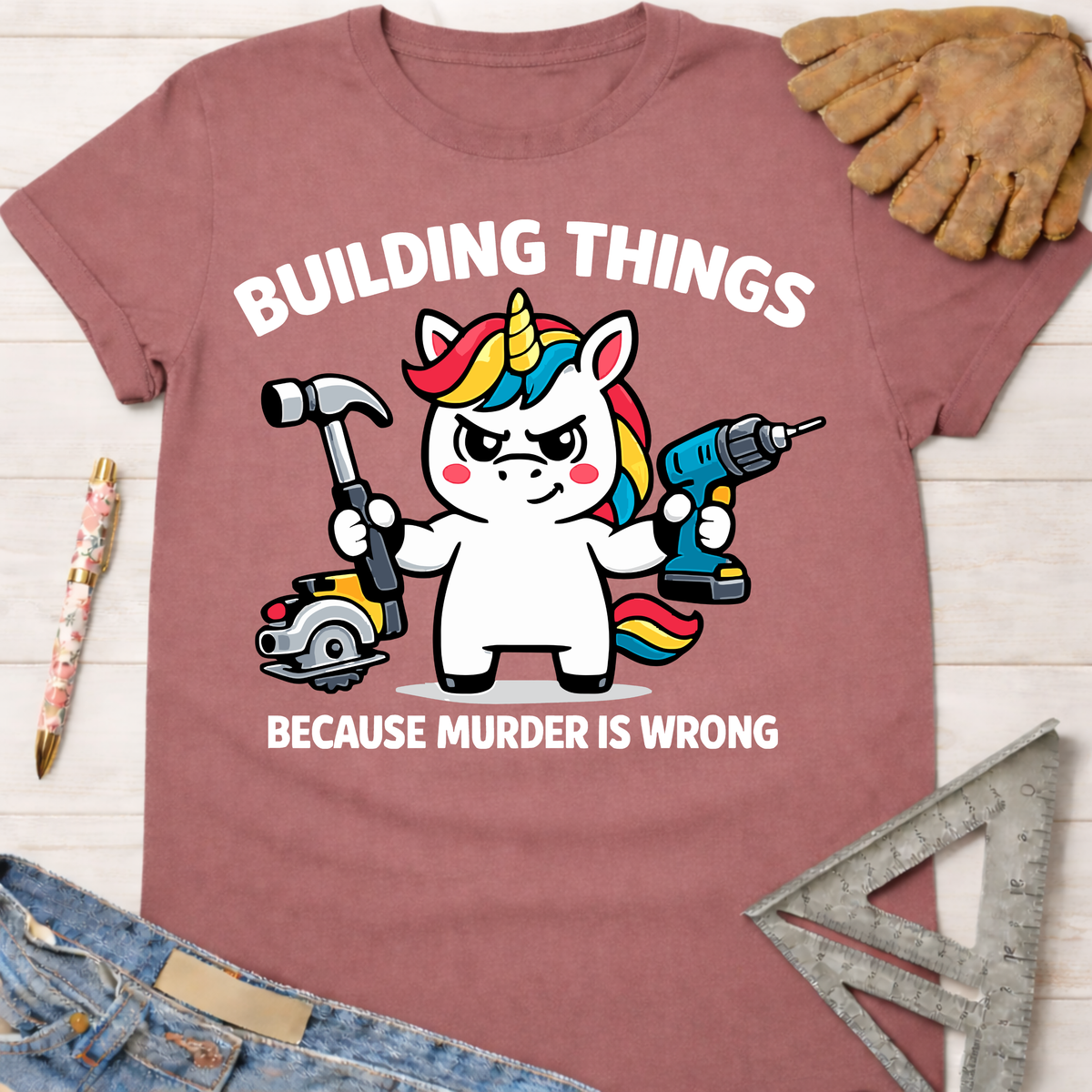 Women's funny woodworking graphic tee shirt for female woodworkers, makers, and DIY builders — Sawdust & Ponytails. Mauve cotton tee with a cartoon unicorn holding a hammer, drill, and saw, text reads "BUILDING THINGS BECAUSE MURDER IS WRONG.