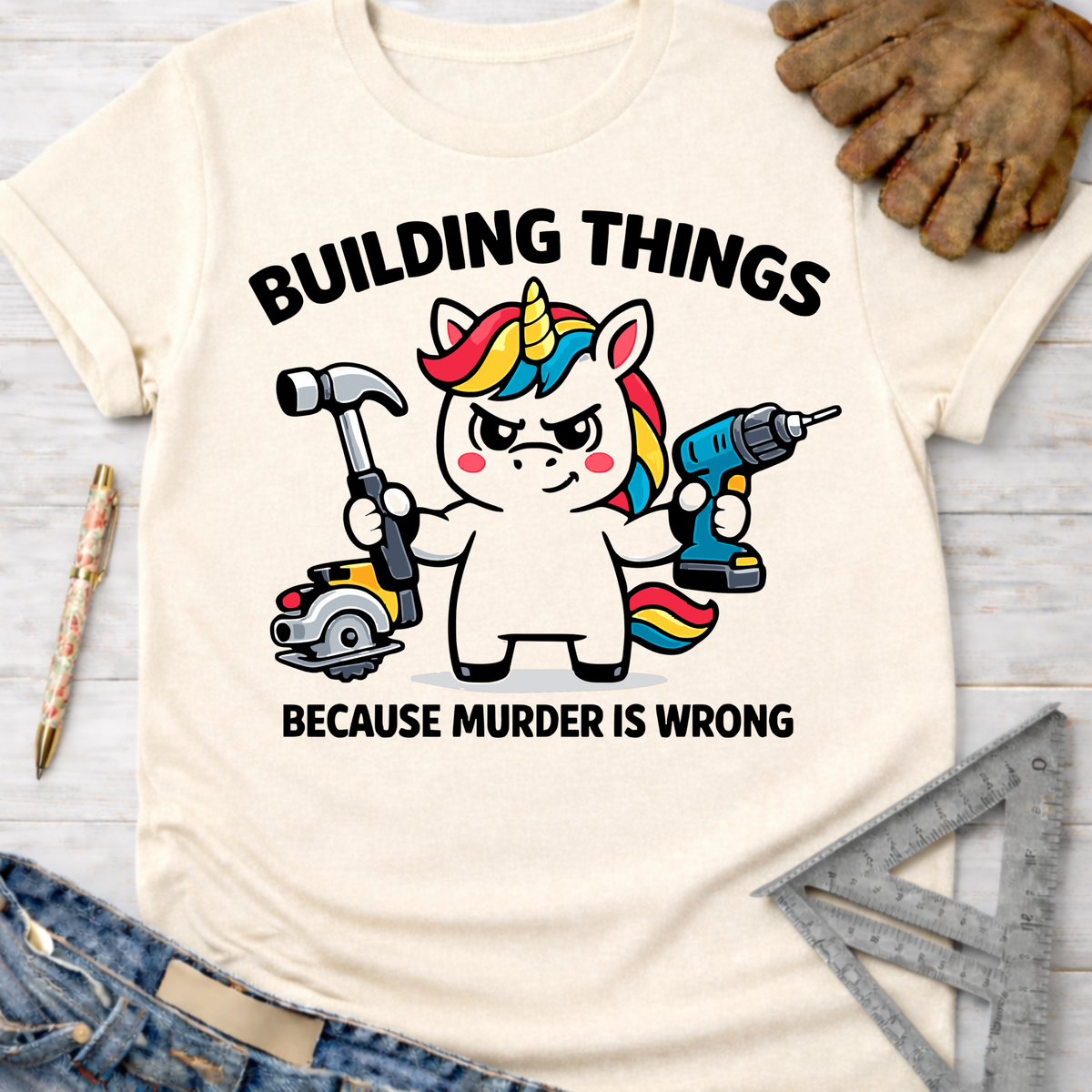 Women's funny woodworking graphic tee shirt for female woodworkers, makers, and DIY builders — Sawdust & Ponytails. Cream t-shirt with a unicorn holding a hammer, circular saw, and drill, text reads "BUILDING THINGS BECAUSE MURDER IS WRONG".