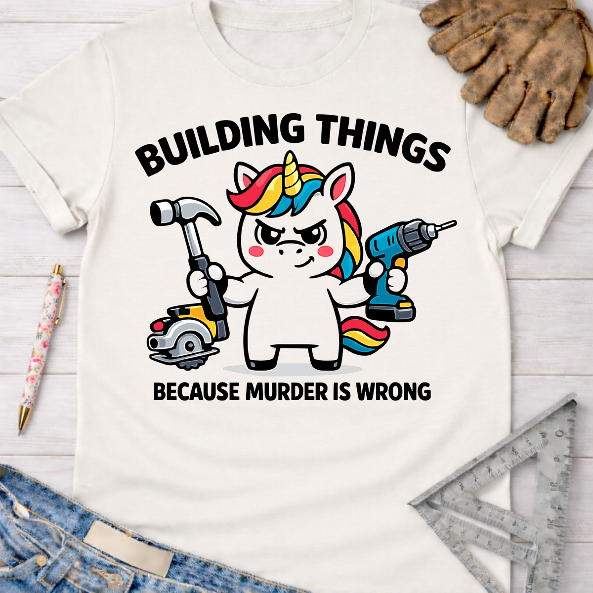 Women's funny woodworking graphic tee shirt for female woodworkers, makers, and DIY builders — Sawdust & Ponytails. White cotton tee with a cartoon unicorn holding a hammer, circular saw, and power drill. Text reads "BUILDING THINGS BECAUSE MURDER IS WRONG.
