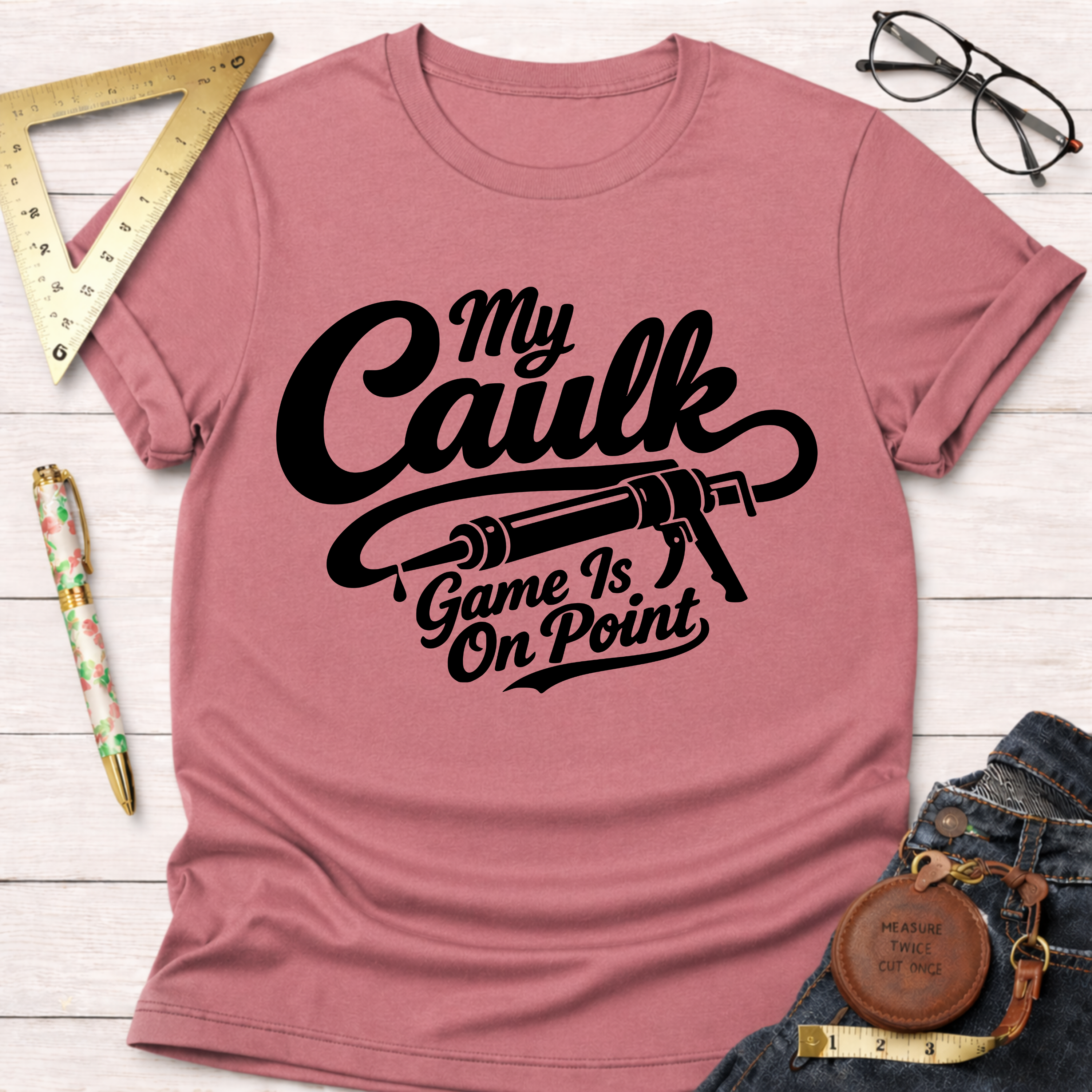 Pink "My Caulk Game Is On Point" t-shirt with a caulk gun graphic, styled with jeans and tools.