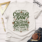 Women's funny woodworking graphic tee shirt for female woodworkers, makers, and DIY builders — Sawdust & Ponytails. Cream t-shirt with "My DIY Playlist: Circular Saw and Creative Swearing on Repeat Until It's Done" text.