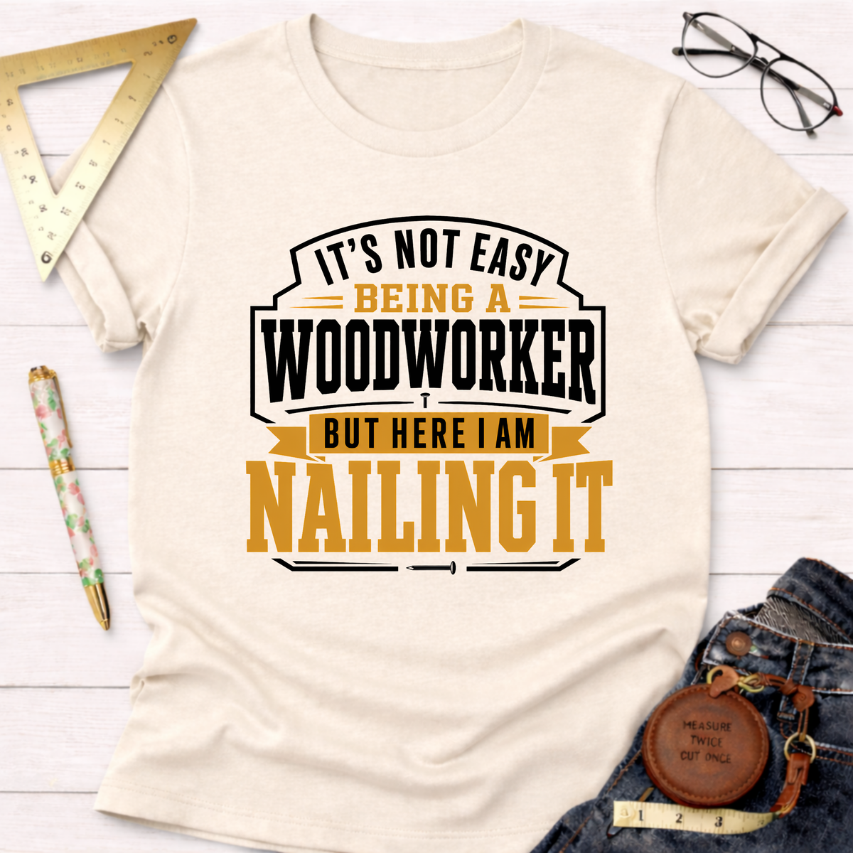 Cream cotton t-shirt with "It's Not Easy Being A Woodworker But Here I Am Nailing It" graphic in black and gold.