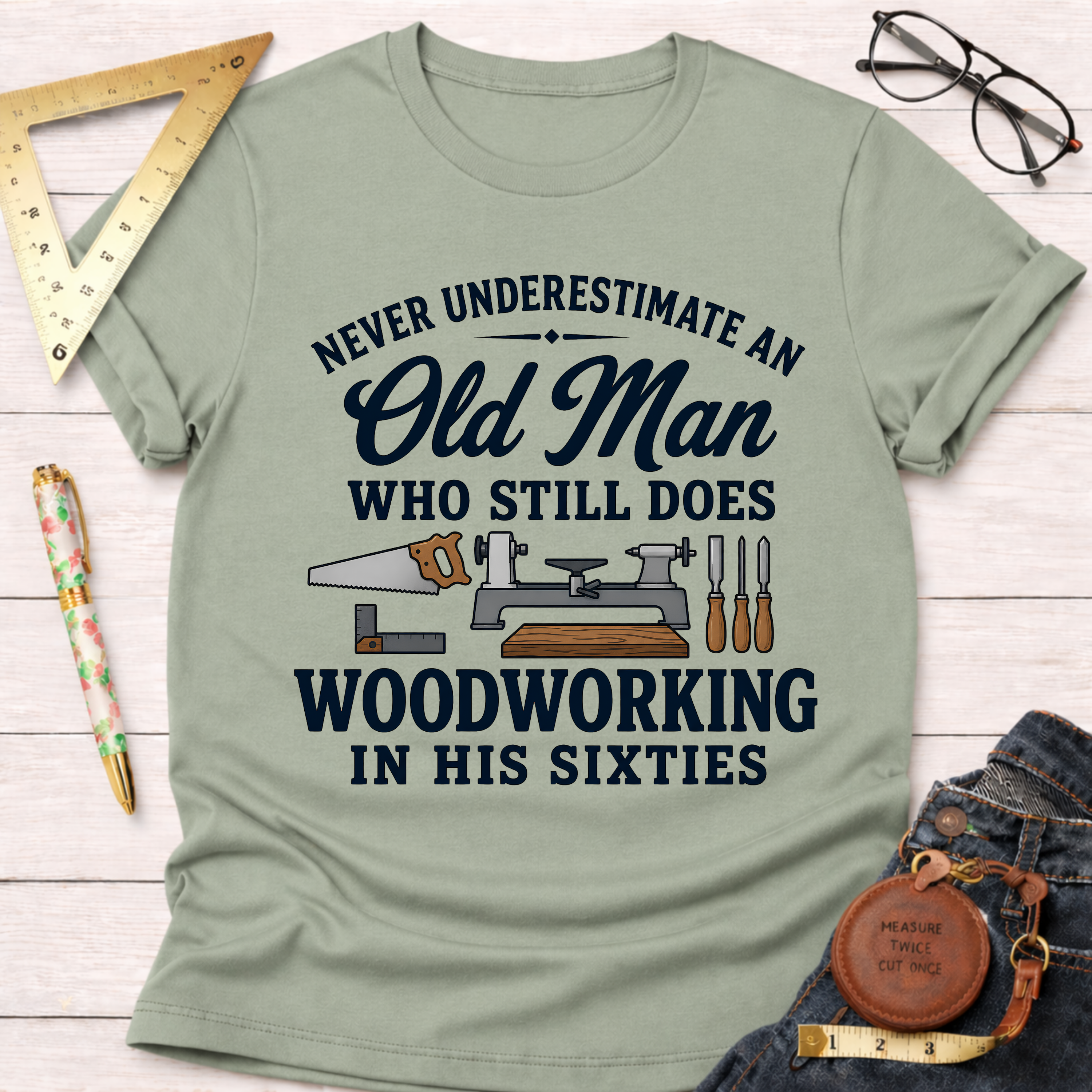 Sage green t-shirt with "Never Underestimate An Old Man Who Still Does Woodworking In His Sixties" text and tools graphic.