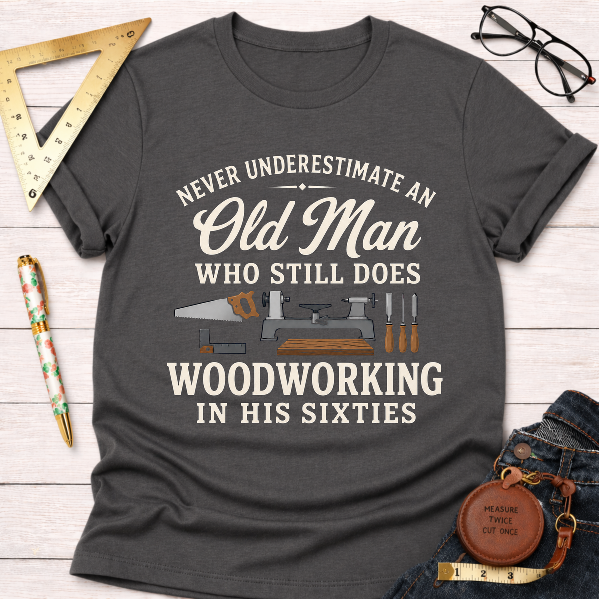 Charcoal gray t-shirt with "Never Underestimate An Old Man Who Still Does Woodworking In His Sixties" text and tools graphic.