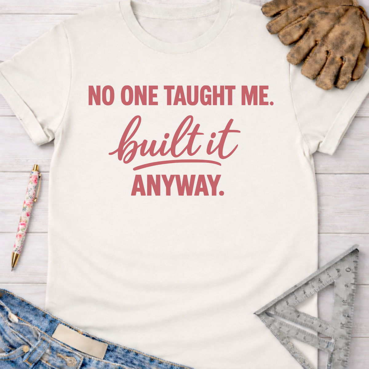 Women's funny woodworking graphic tee shirt for female woodworkers, makers, and DIY builders — Sawdust & Ponytails. Cream t-shirt with "NO ONE TAUGHT ME. built it ANYWAY." in pink text, styled with jeans, work gloves, and a ruler.