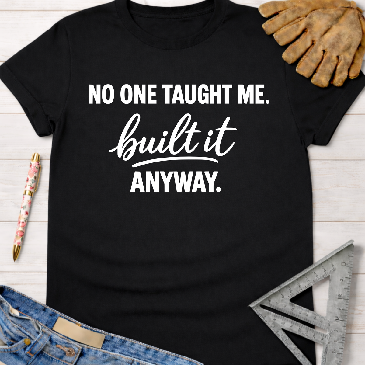 Black cotton "No One Taught Me. Built It Anyway." graphic tee with work glove, pen, ruler, and jeans. Women's funny woodworking graphic tee shirt for female woodworkers, makers, and DIY builders — Sawdust & Ponytails