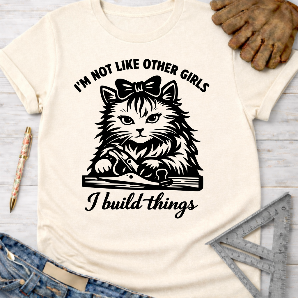 Women's funny woodworking graphic tee shirt for female woodworkers, makers, and DIY builders — Sawdust & Ponytails. Cream t-shirt with a black graphic of a cat with a bow, using a hand plane, and text "I'm not like other girls, I build things.