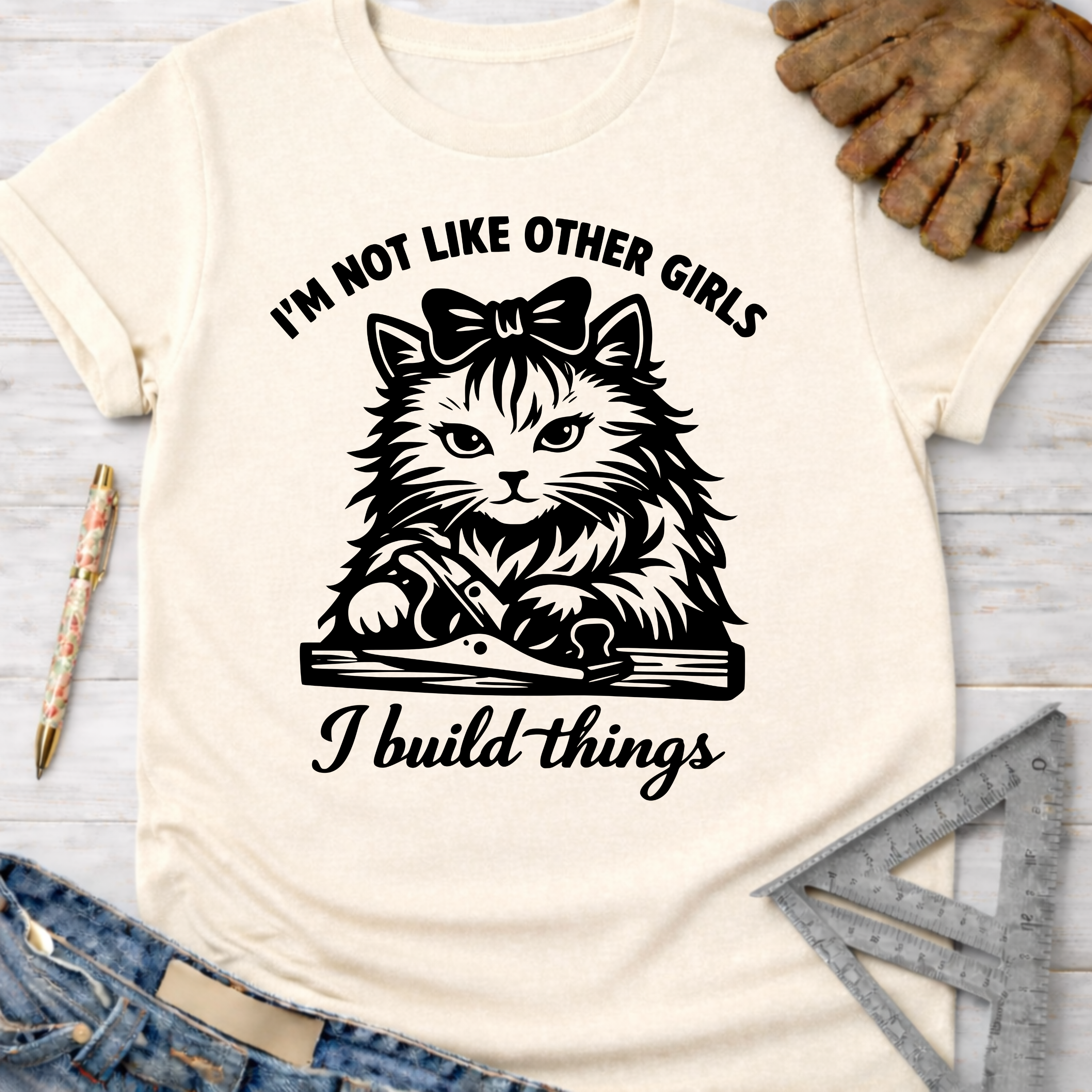Women's funny woodworking graphic tee shirt for female woodworkers, makers, and DIY builders — Sawdust & Ponytails. Cream t-shirt with a black graphic of a cat with a bow, using a hand plane, and text "I'm not like other girls, I build things.