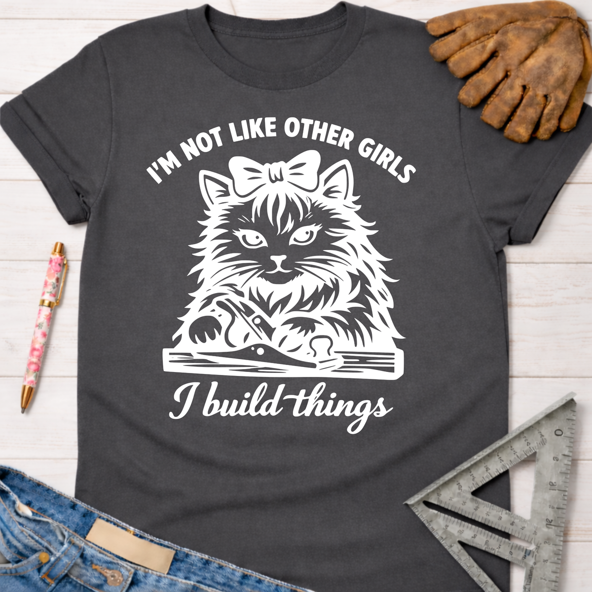 Women's funny woodworking graphic tee shirt for female woodworkers, makers, and DIY builders — Sawdust & Ponytails. Dark gray t-shirt with a white graphic of a cat with a bow, holding a hand plane, and text "I'm not like other girls I build things".