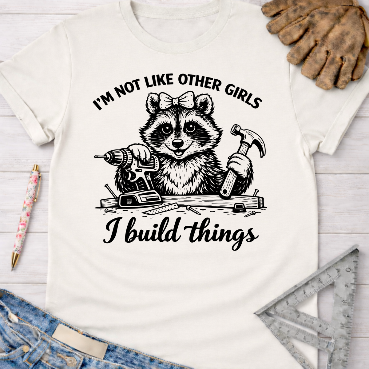 Women's funny woodworking graphic tee shirt for female woodworkers, makers, and DIY builders — Sawdust & Ponytails. White cotton tee with a raccoon holding a drill and hammer, text "I'm not like other girls I build things.