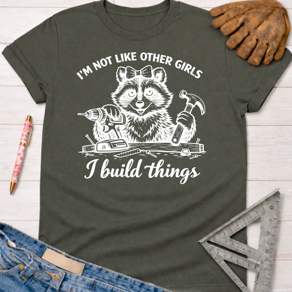 Women's funny woodworking graphic tee shirt for female woodworkers, makers, and DIY builders — Sawdust & Ponytails. Dark gray tee with a raccoon holding a drill and hammer, text "I'm not like other girls I build things.