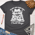 Women's funny woodworking graphic tee shirt for female woodworkers, makers, and DIY builders — Sawdust & Ponytails. Dark gray tee with white "I'm not like other girls, I build things" text and a Highland cow with a bow and wood plane.