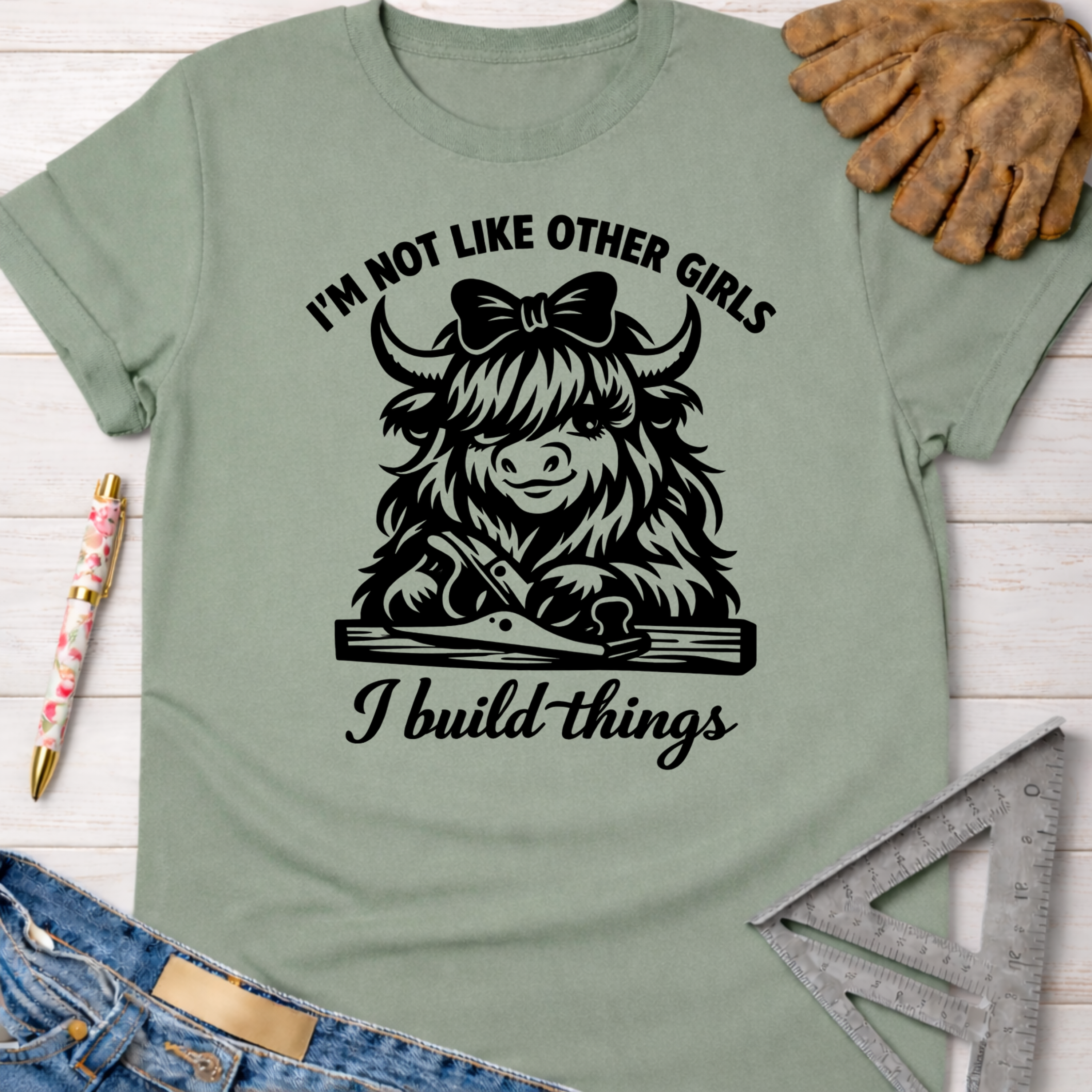 Women's funny woodworking graphic tee shirt for female woodworkers, makers, and DIY builders — Sawdust & Ponytails. Sage green cotton tee with a black graphic of a Highland cow wearing a bow, holding a hand plane, and text "I'm not like other girls I build things.