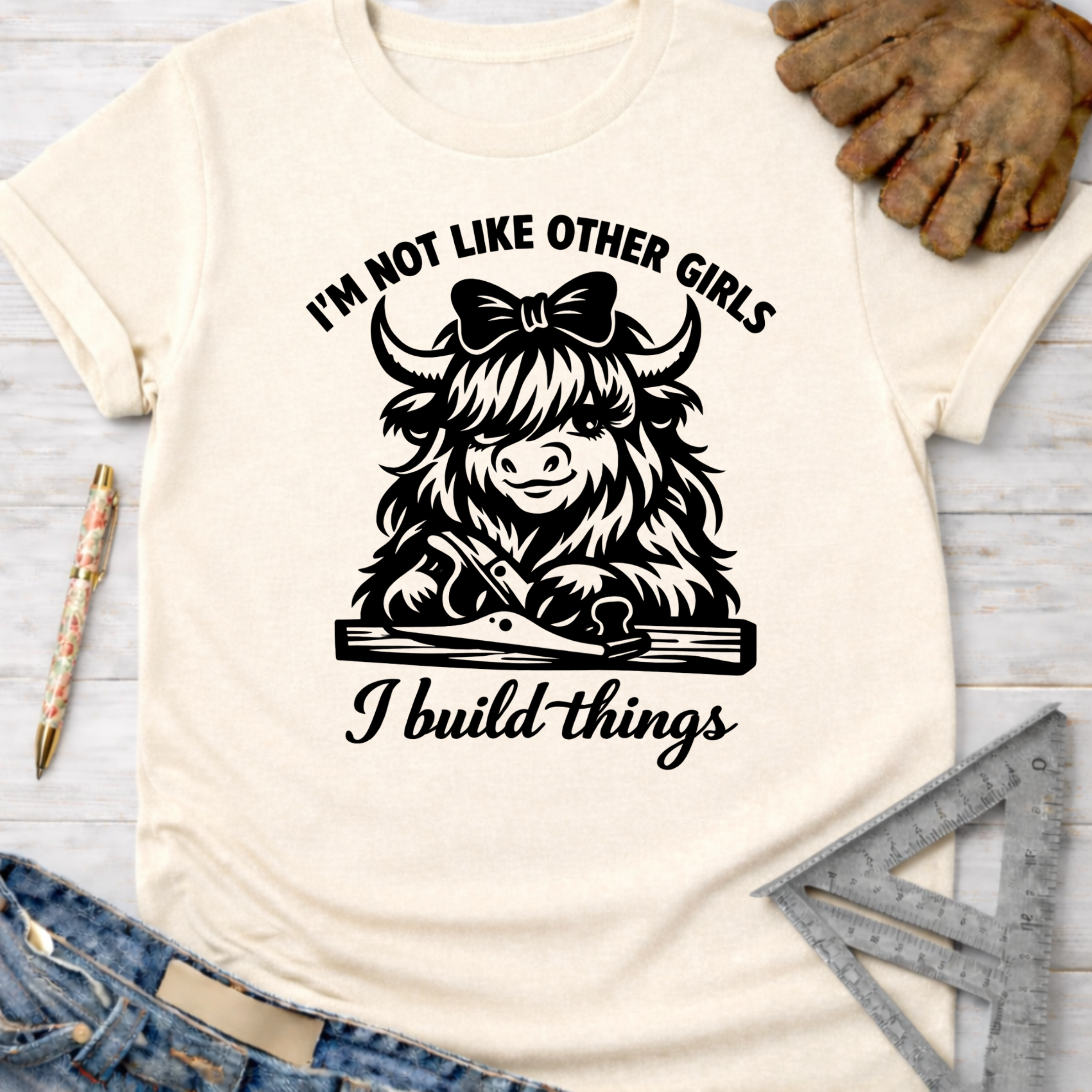 Women's funny woodworking graphic tee shirt for female woodworkers, makers, and DIY builders — Sawdust & Ponytails. Cream cotton tee with a black graphic of a Highland cow using a hand plane, text "I'm not like other girls I build things".