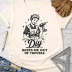 Women's funny woodworking graphic tee shirt for female woodworkers, makers, and DIY builders — Sawdust & Ponytails. Cream t-shirt with a retro woman holding a drill and tool belt, text "DIY KEEPS ME OUT OF TROUBLE.