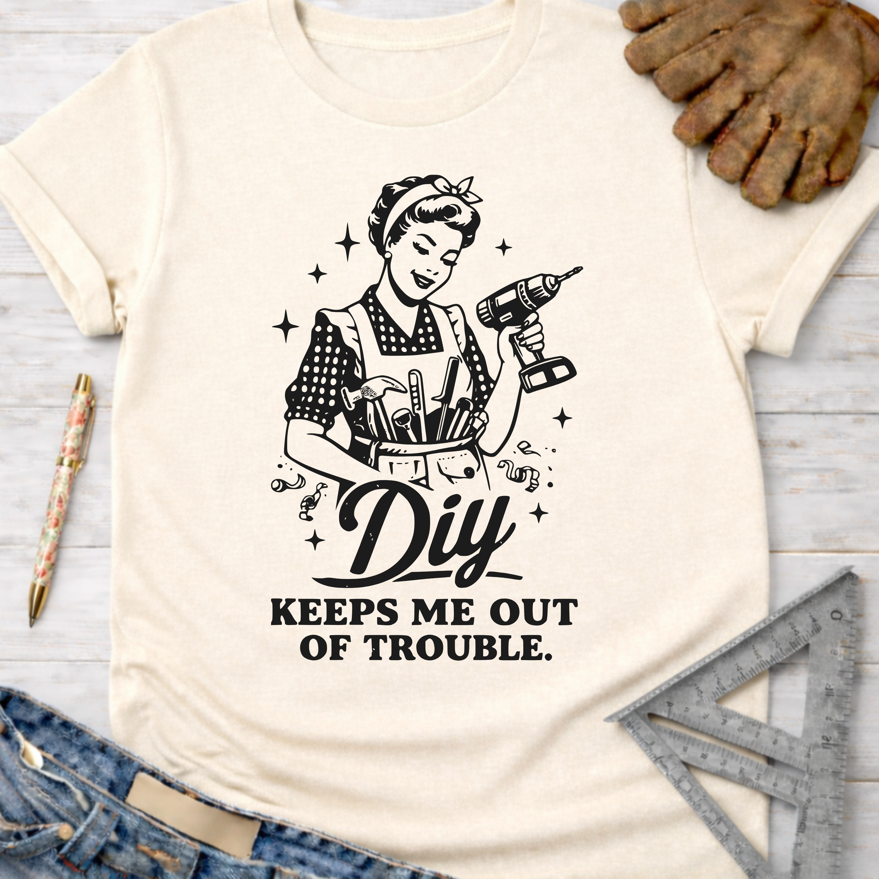 Women's funny woodworking graphic tee shirt for female woodworkers, makers, and DIY builders — Sawdust & Ponytails. Cream t-shirt with a retro woman holding a drill and tool belt, text "DIY KEEPS ME OUT OF TROUBLE.
