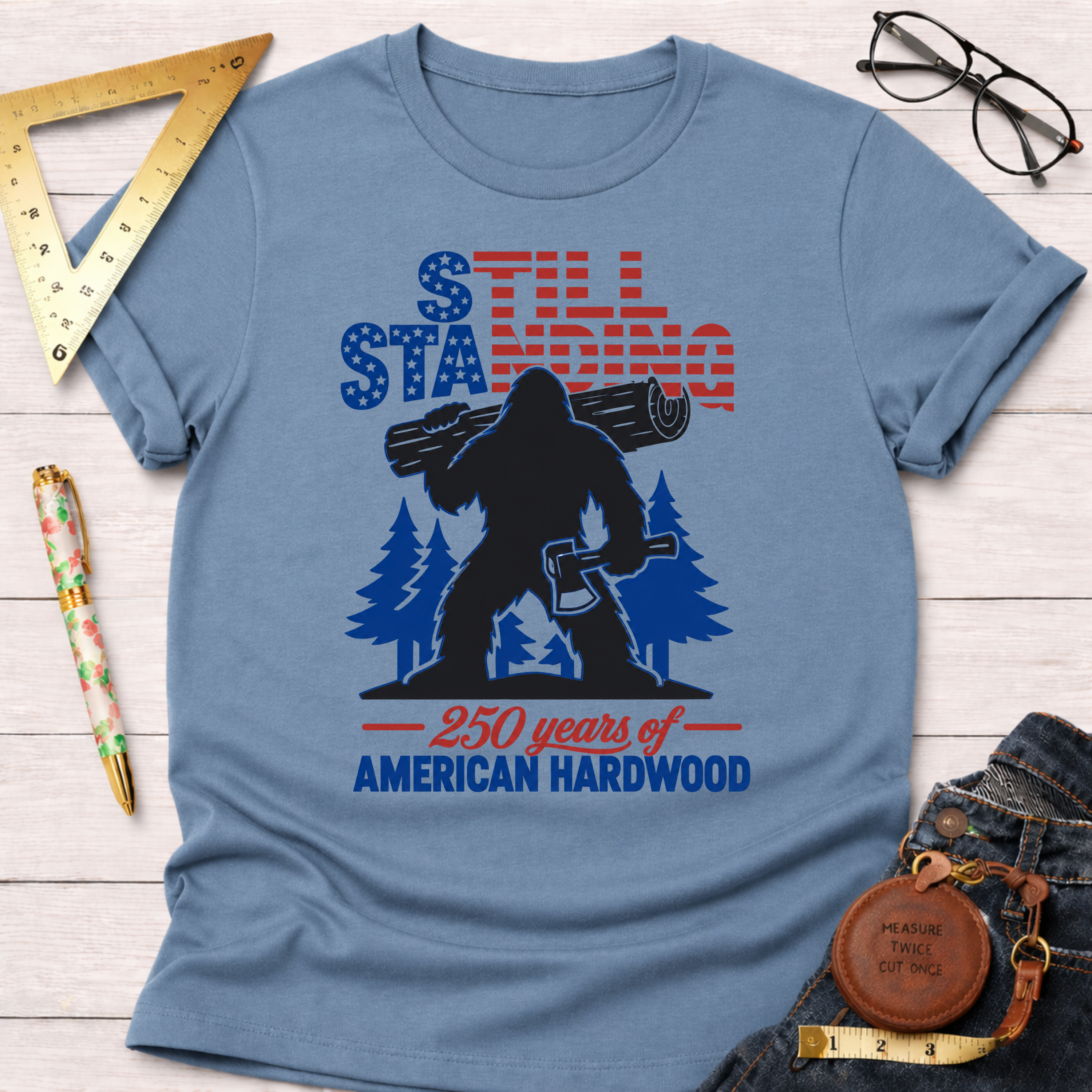 Light blue cotton t-shirt with a patriotic Sasquatch holding an axe and log, "Still Standing 250 years of American Hardwood" text.