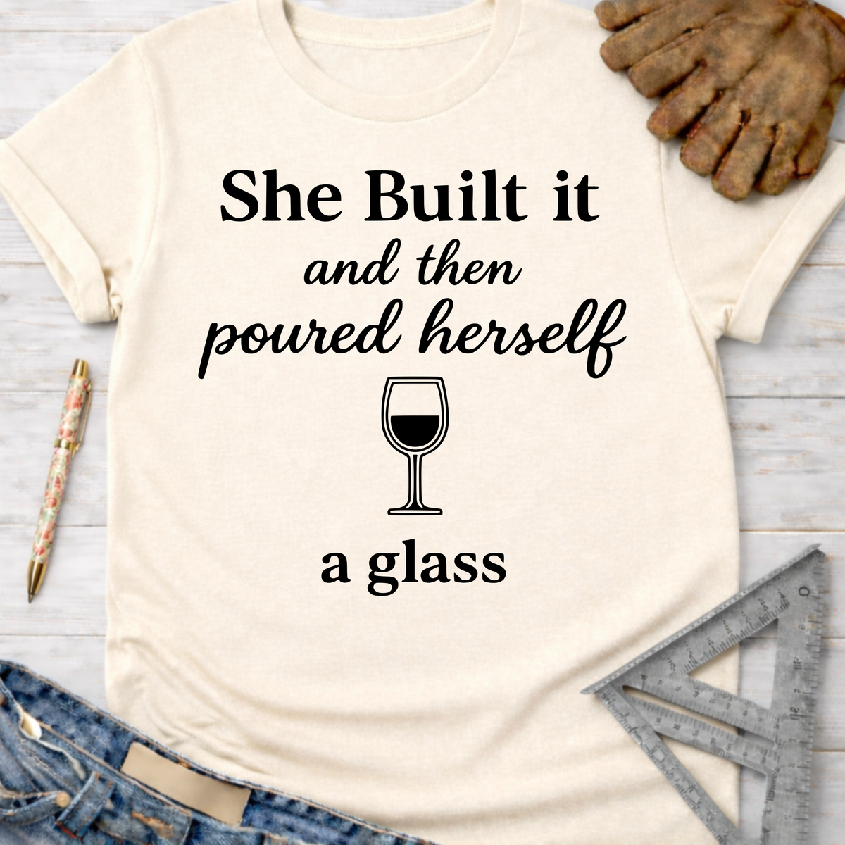 Women's funny woodworking graphic tee shirt for female woodworkers, makers, and DIY builders — Sawdust & Ponytails. Cream t-shirt with "She Built it and then poured herself a glass" text and wine glass graphic.