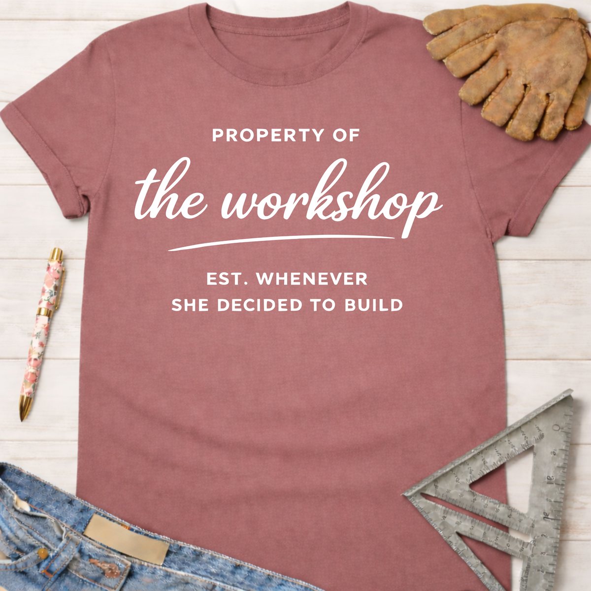 Women's funny woodworking graphic tee shirt for female woodworkers, makers, and DIY builders — Sawdust & Ponytails. Mauve t-shirt with "Property of the workshop EST. WHENEVER SHE DECIDED TO BUILD" in white text.