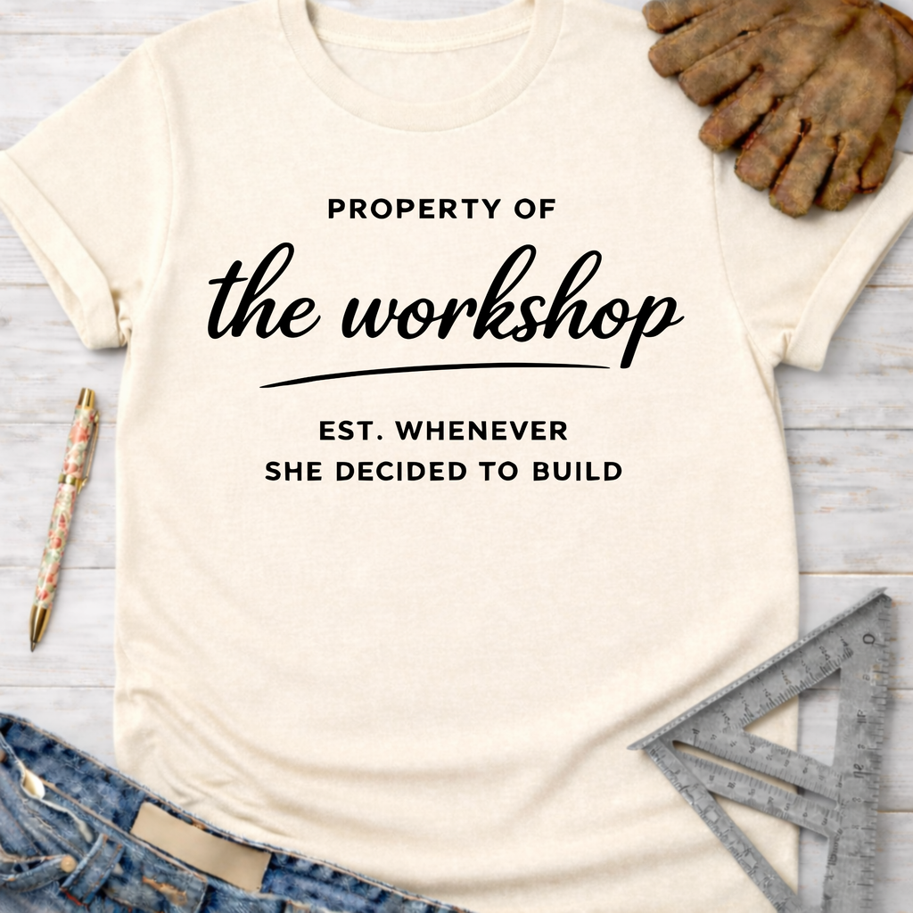 Women's funny woodworking graphic tee shirt for female woodworkers, makers, and DIY builders — Sawdust & Ponytails. Cream t-shirt with "Property of the workshop Est. Whenever She Decided to Build" in black text, styled with jeans, work glove, and ruler.
