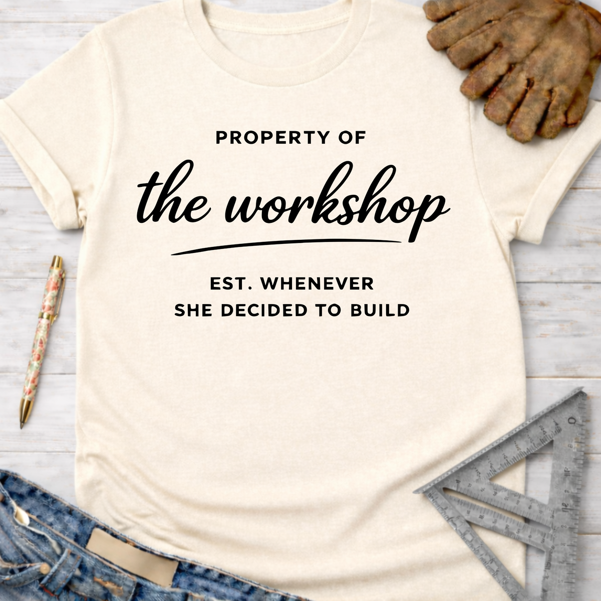 Women's funny woodworking graphic tee shirt for female woodworkers, makers, and DIY builders — Sawdust & Ponytails. Cream t-shirt with "Property of the workshop Est. Whenever She Decided to Build" in black text, styled with jeans, work glove, and ruler.