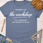 Women's funny woodworking graphic tee shirt for female woodworkers, makers, and DIY builders — Sawdust & Ponytails. Blue cotton t-shirt with "Property of the workshop EST. Whenever She Decided to Build" in white text.
