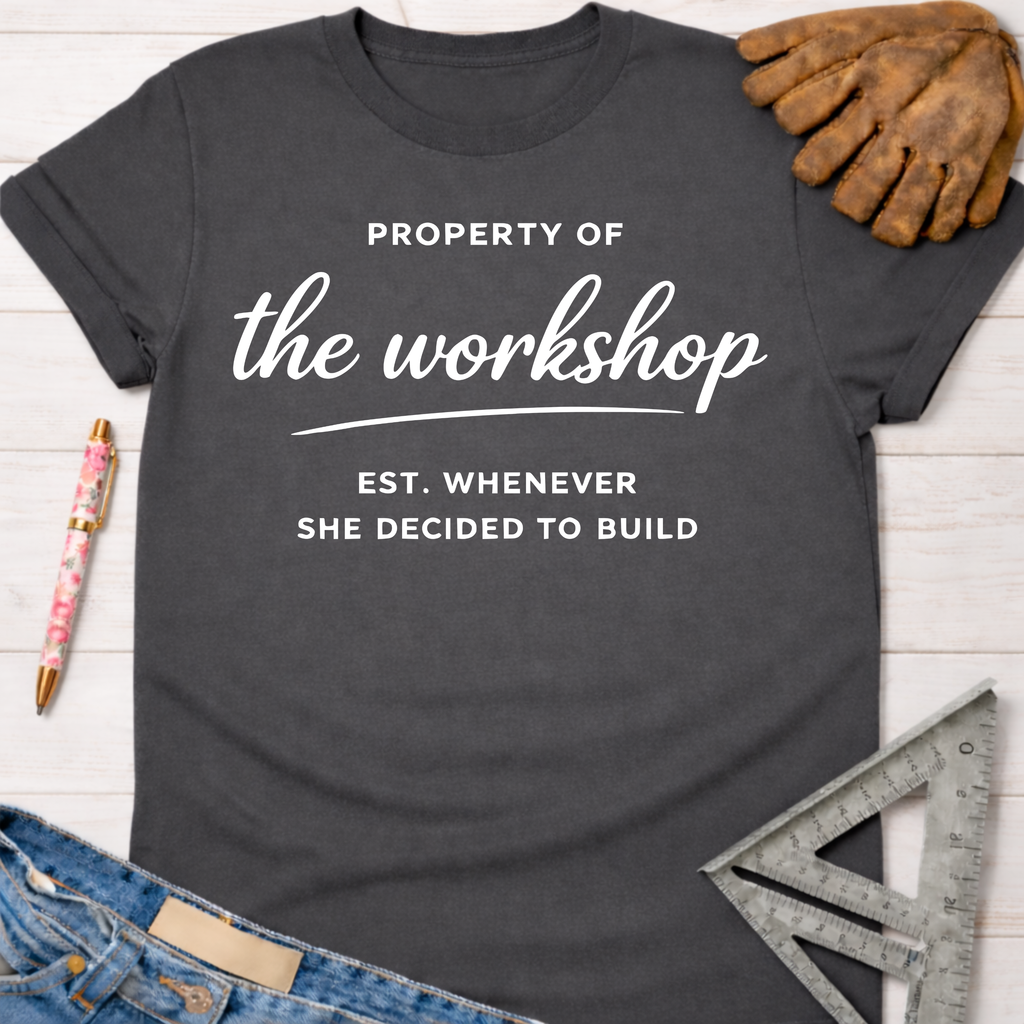 Women's funny woodworking graphic tee shirt for female woodworkers, makers, and DIY builders — Sawdust & Ponytails. Dark gray cotton tee with "Property of the workshop EST. WHENEVER SHE DECIDED TO BUILD" in white text.
