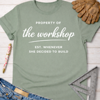 Women's funny woodworking graphic tee shirt for female woodworkers, makers, and DIY builders — Sawdust & Ponytails. Sage green cotton t-shirt with "Property of the workshop EST. WHENEVER SHE DECIDED TO BUILD" in white text.