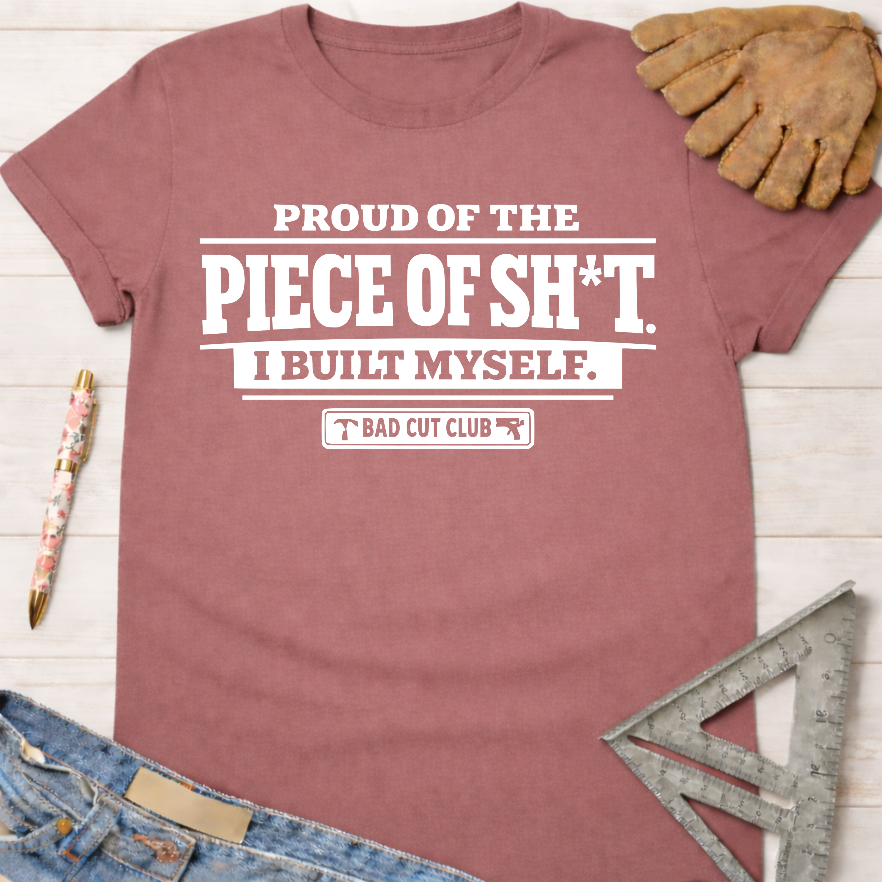 Women's funny woodworking graphic tee shirt for female woodworkers, makers, and DIY builders — Sawdust & Ponytails. Mauve cotton tee with "PROUD OF THE PIECE OF SH*T. I BUILT MYSELF. BAD CUT CLUB" in white text.