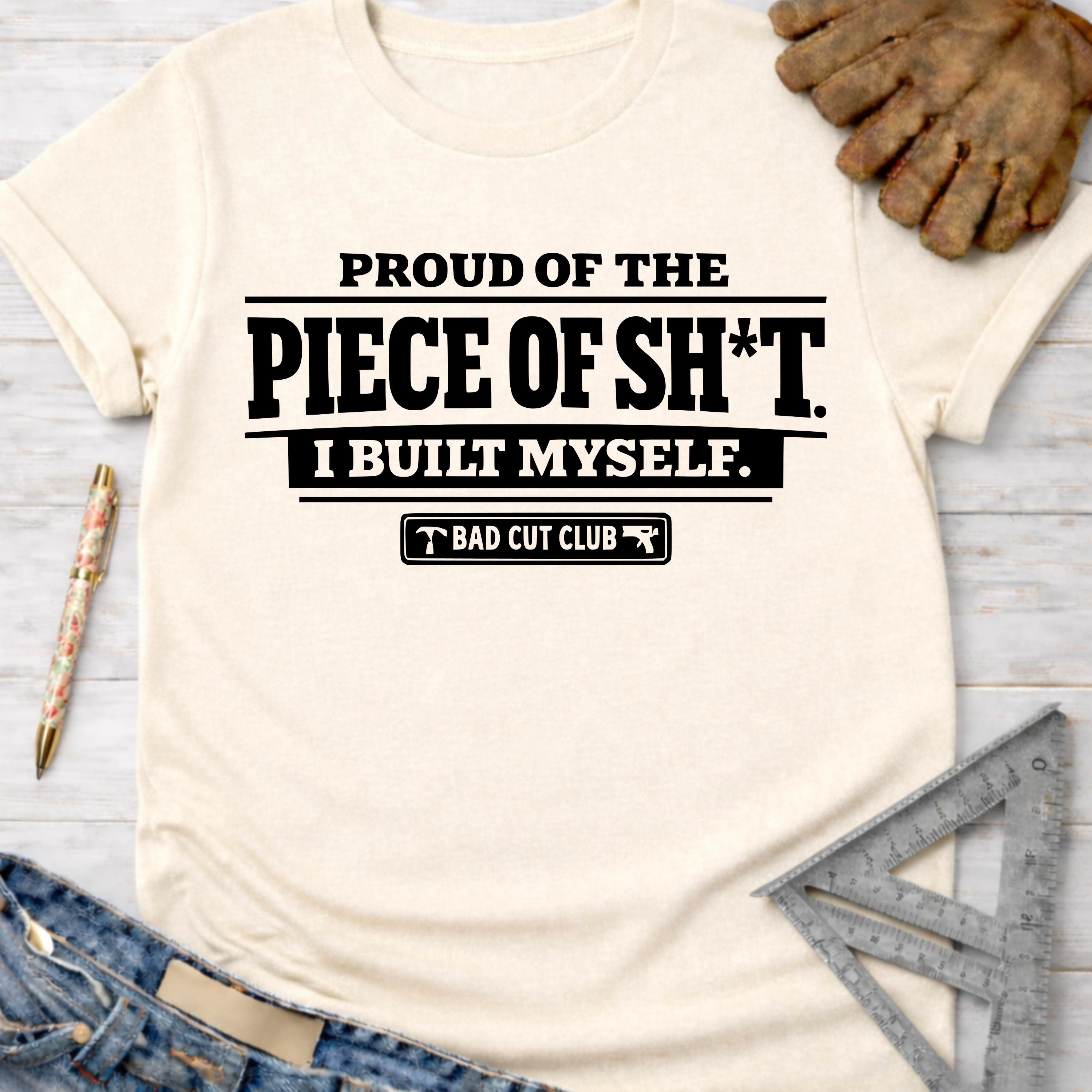Women's funny woodworking graphic tee shirt for female woodworkers, makers, and DIY builders — Sawdust & Ponytails. Cream t-shirt with "Proud of the Piece of Sh*t I Built Myself. Bad Cut Club" text.