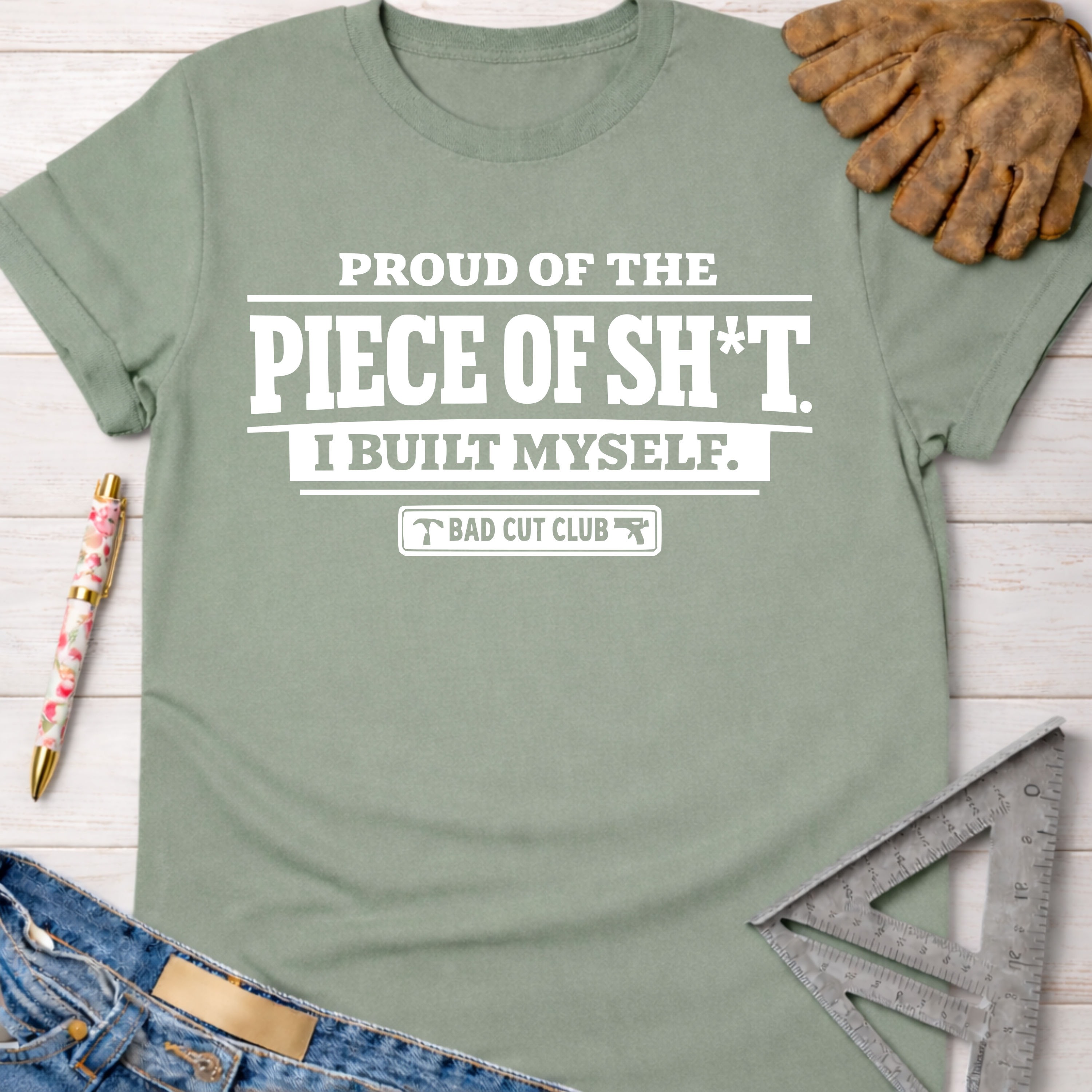 Sage green women's funny woodworking graphic tee with "PROUD OF THE PIECE OF SH*T. I BUILT MYSELF. BAD CUT CLUB" text.