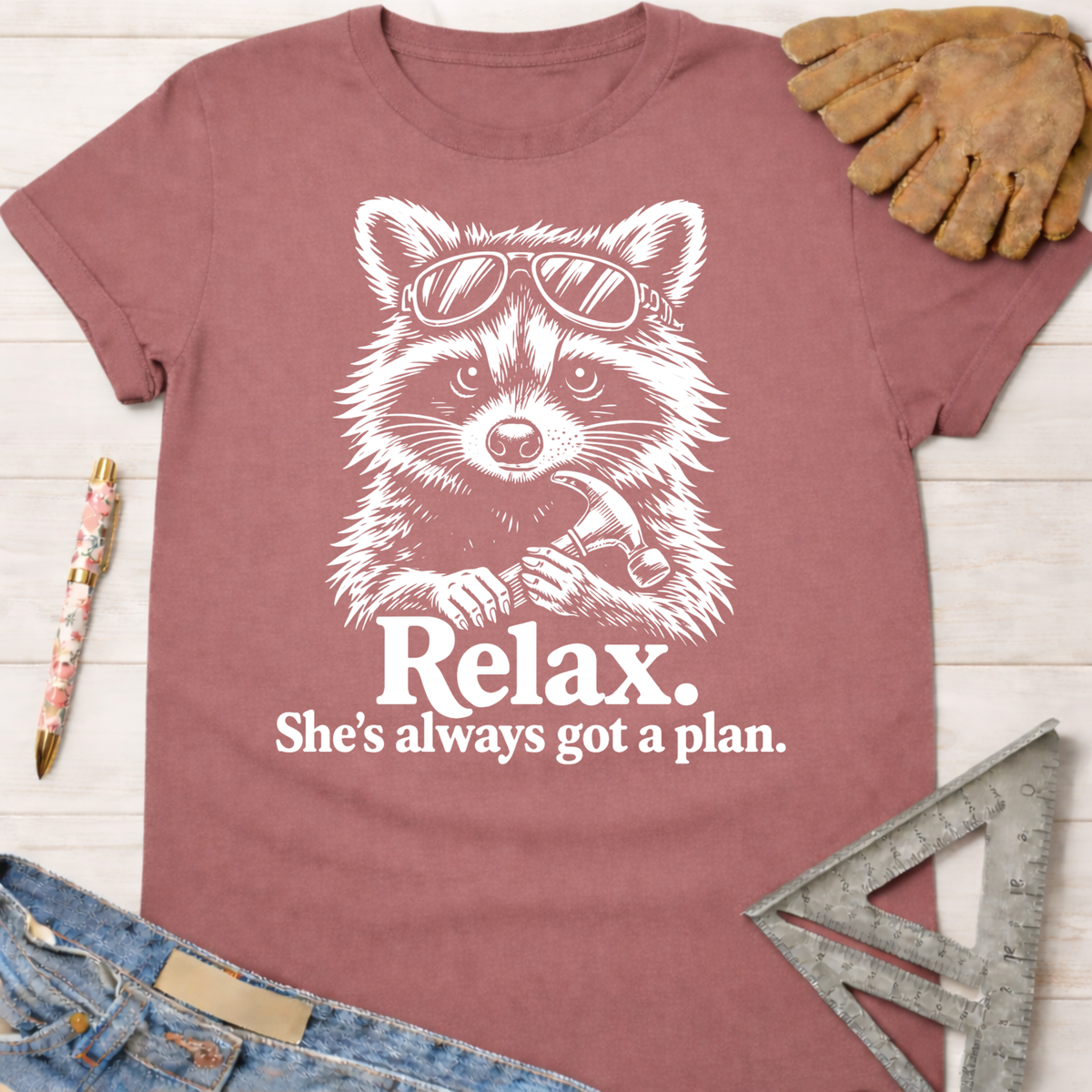 Women's funny woodworking graphic tee shirt for female woodworkers, makers, and DIY builders — Sawdust & Ponytails. Mauve t-shirt with a white graphic of a raccoon wearing sunglasses and holding a hammer, with text "Relax. She's always got a plan.