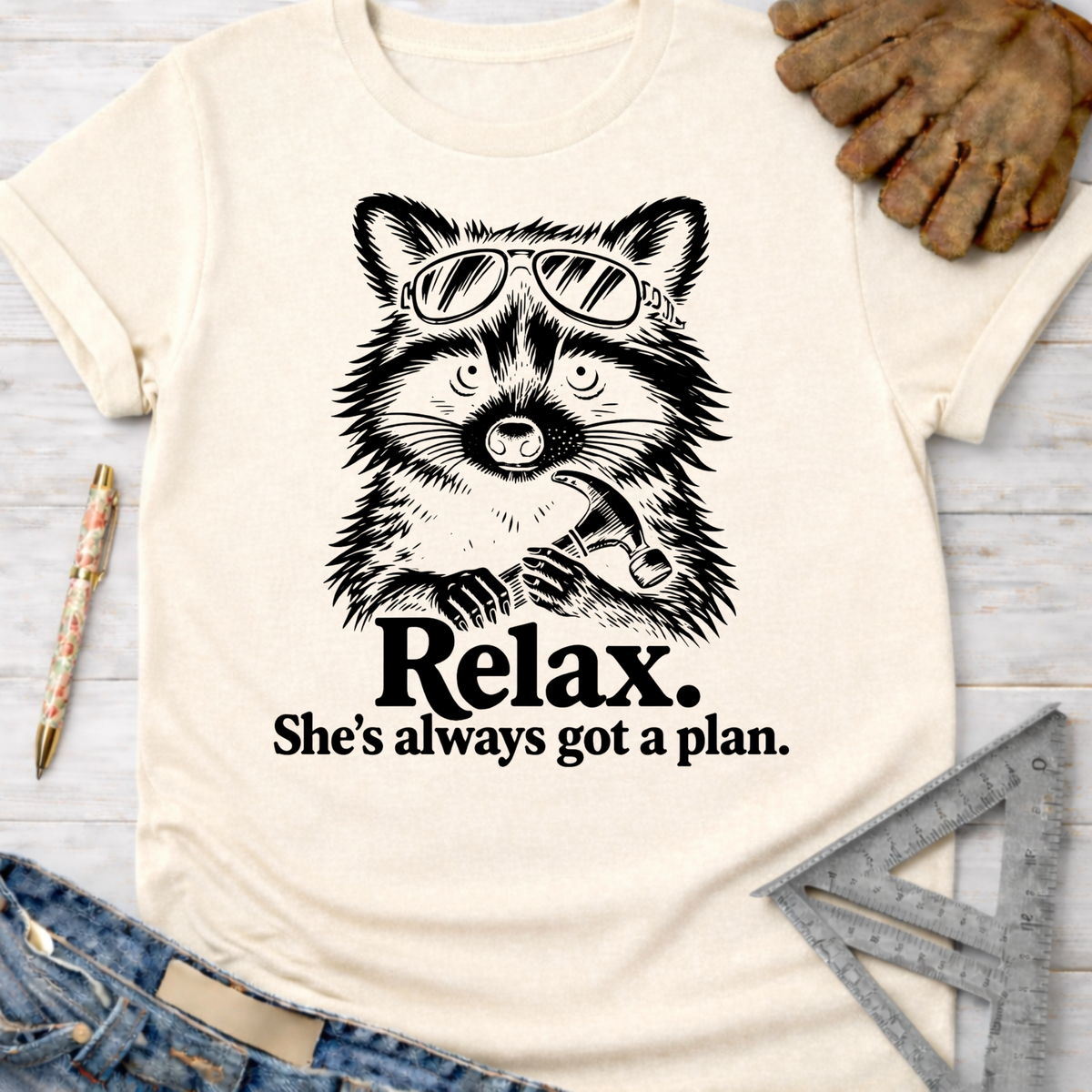 Women's funny woodworking graphic tee shirt for female woodworkers, makers, and DIY builders — Sawdust & Ponytails. Cream t-shirt with a black graphic of a raccoon wearing sunglasses and holding a hammer, with the text "Relax. She's always got a plan.