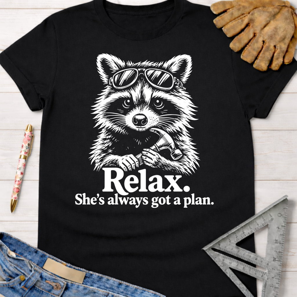 Black cotton tee with a white graphic of a raccoon in sunglasses holding a hammer, text "Relax. She's always got a plan." Women's funny woodworking graphic tee shirt for female woodworkers, makers, and DIY builders — Sawdust & Ponytails.