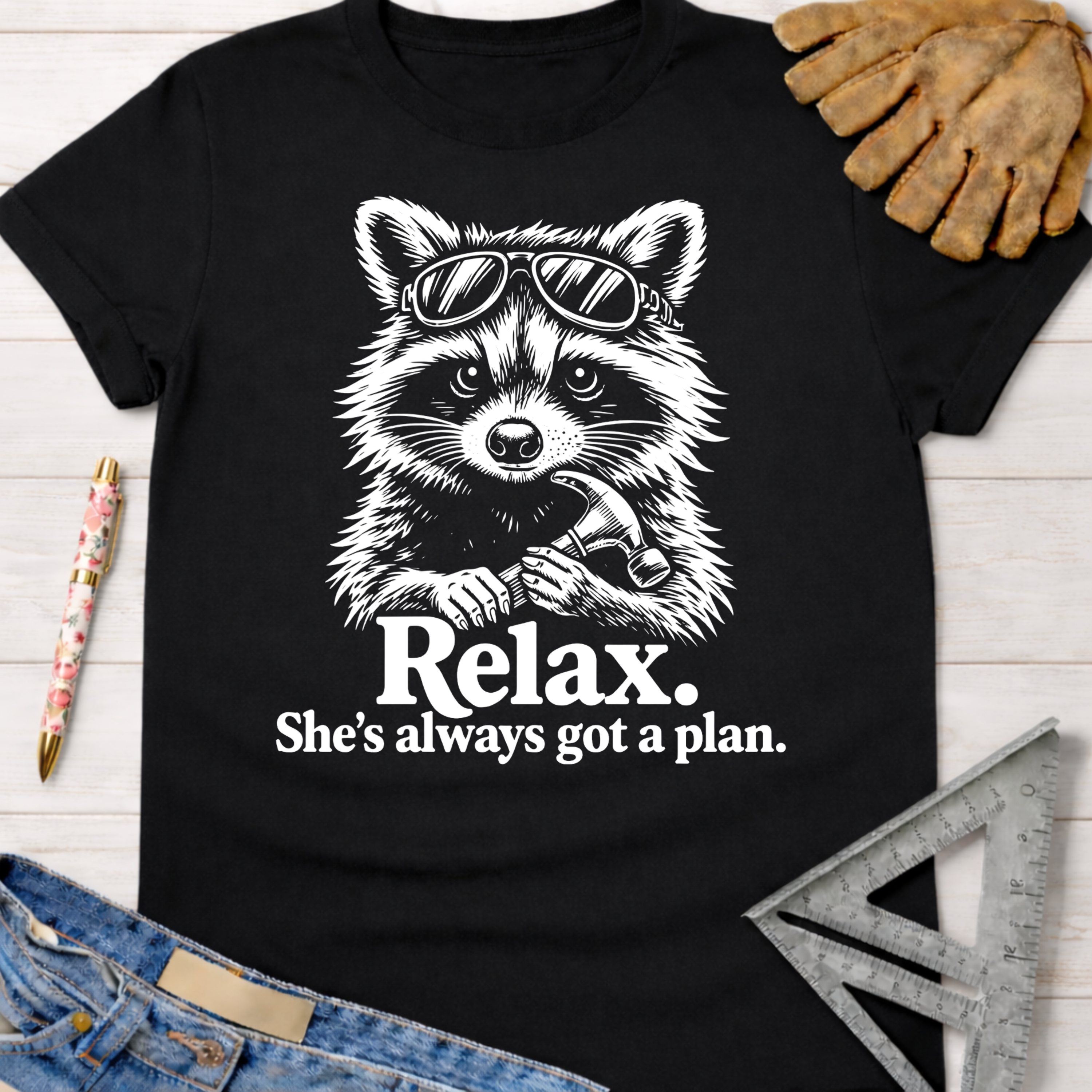 Black cotton tee with a white graphic of a raccoon in sunglasses holding a hammer, text "Relax. She's always got a plan." Women's funny woodworking graphic tee shirt for female woodworkers, makers, and DIY builders — Sawdust & Ponytails.