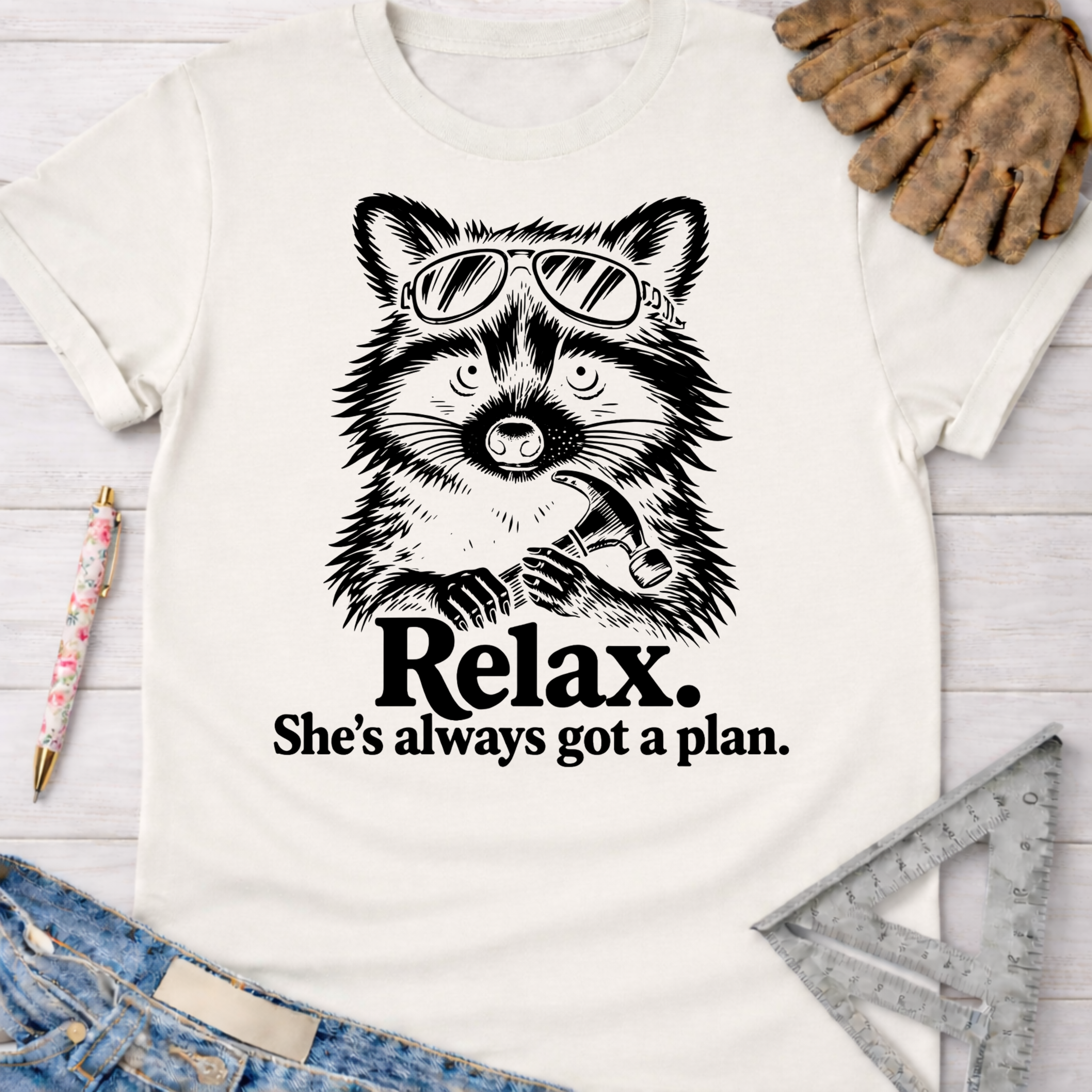 Women's funny woodworking graphic tee shirt for female woodworkers, makers, and DIY builders — Sawdust & Ponytails. White cotton tee with a black graphic of a raccoon wearing sunglasses and holding a hammer, with text "Relax. She's always got a plan.
