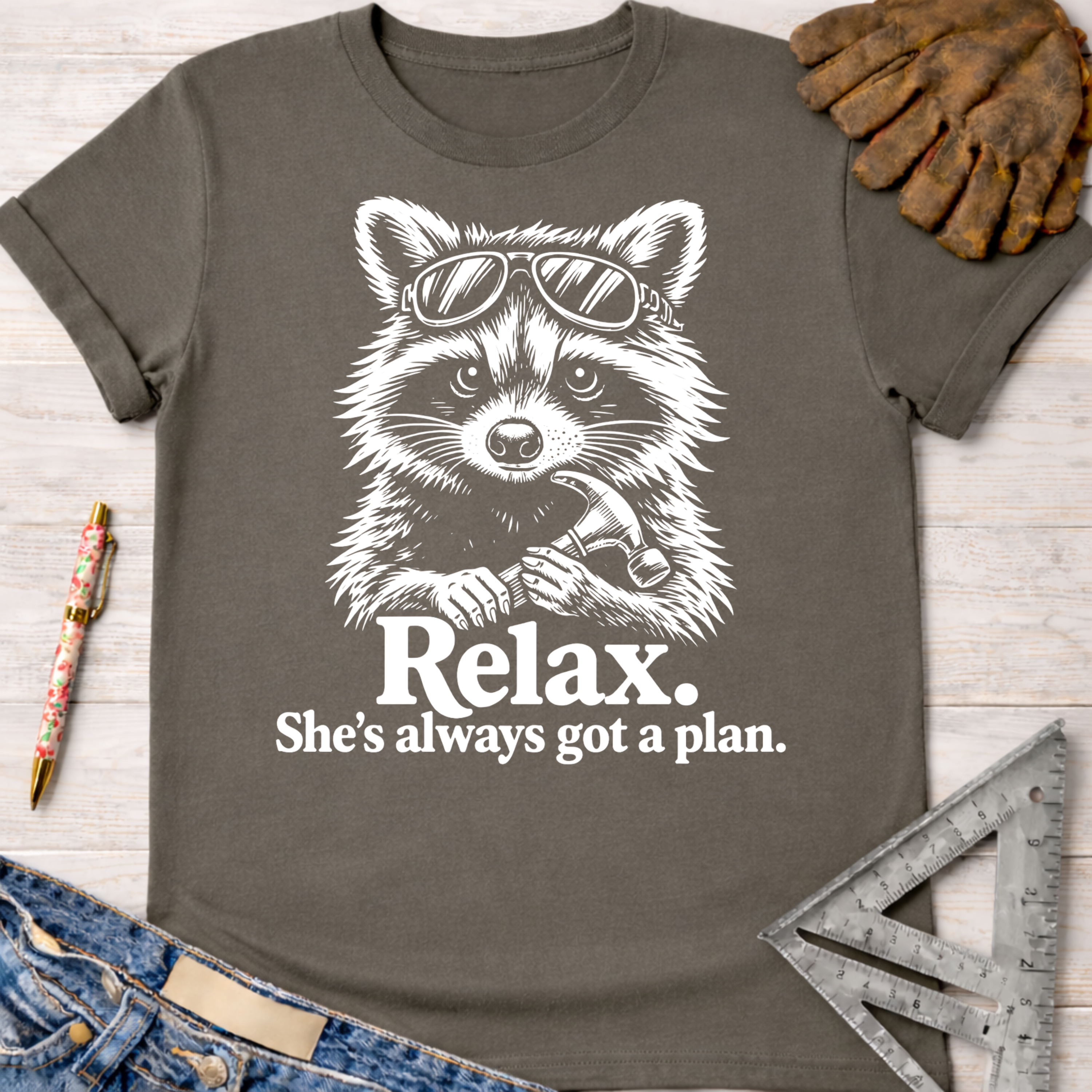 Women's funny woodworking graphic tee shirt for female woodworkers, makers, and DIY builders — Sawdust & Ponytails. Dark gray t-shirt with a white graphic of a raccoon wearing sunglasses and holding a hammer, with "Relax. She's always got a plan." text.