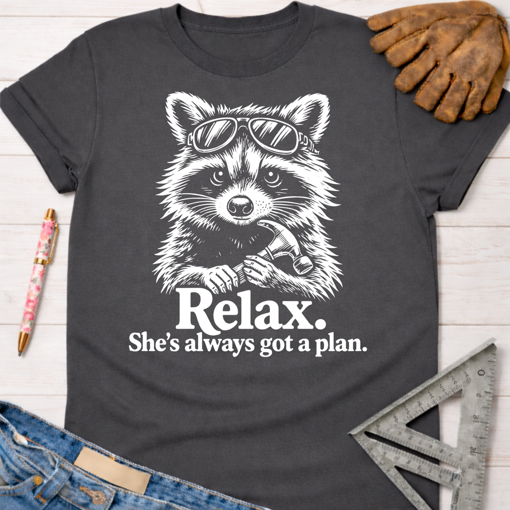 Women's funny woodworking graphic tee shirt for female woodworkers, makers, and DIY builders — Sawdust & Ponytails. Dark gray cotton tee with a white graphic of a raccoon wearing sunglasses and holding a hammer, with the text "Relax. She's always got a plan.