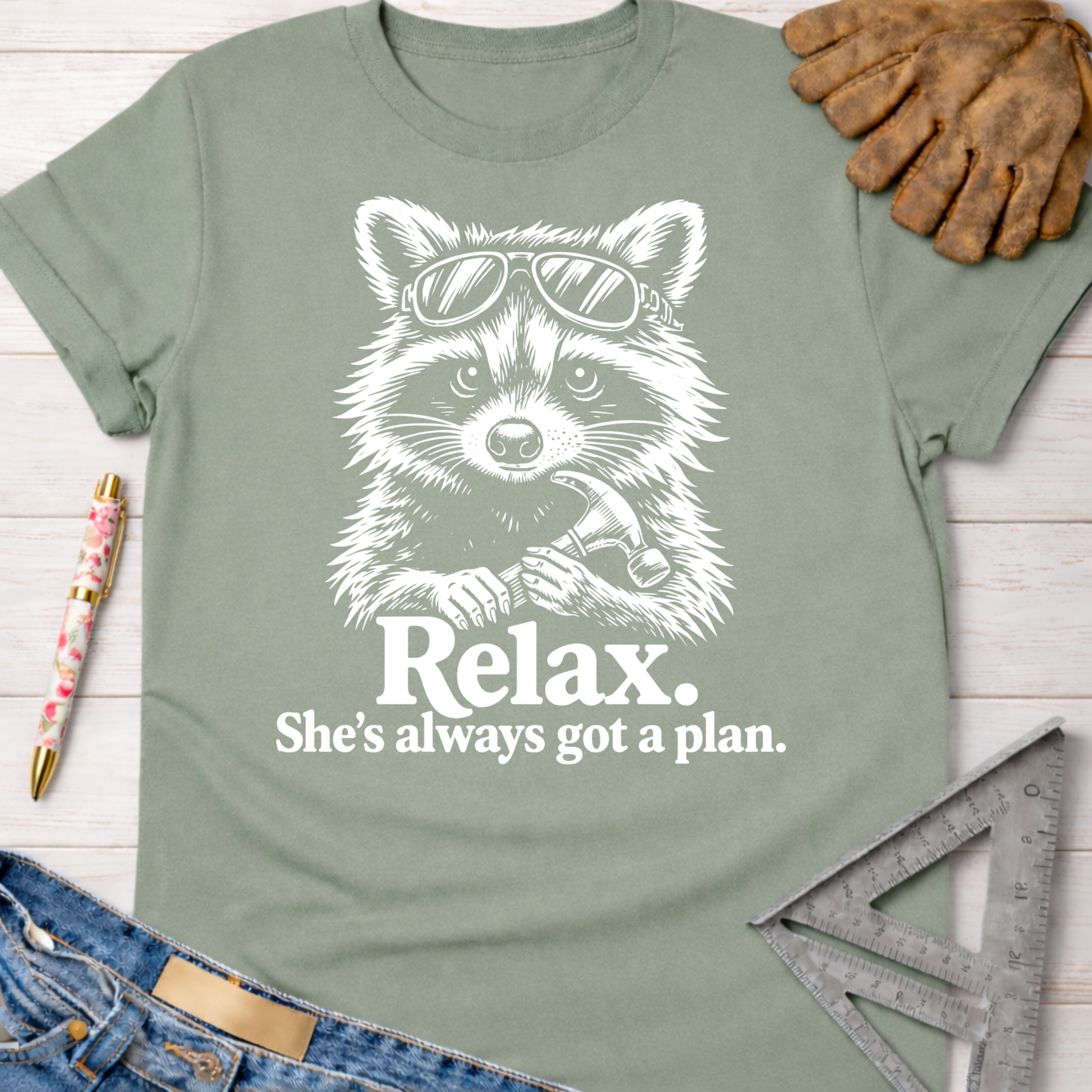 Women's funny woodworking graphic tee shirt for female woodworkers, makers, and DIY builders — Sawdust & Ponytails. Sage green cotton tee with a white graphic of a raccoon in sunglasses holding a hammer, with text "Relax. She's always got a plan.