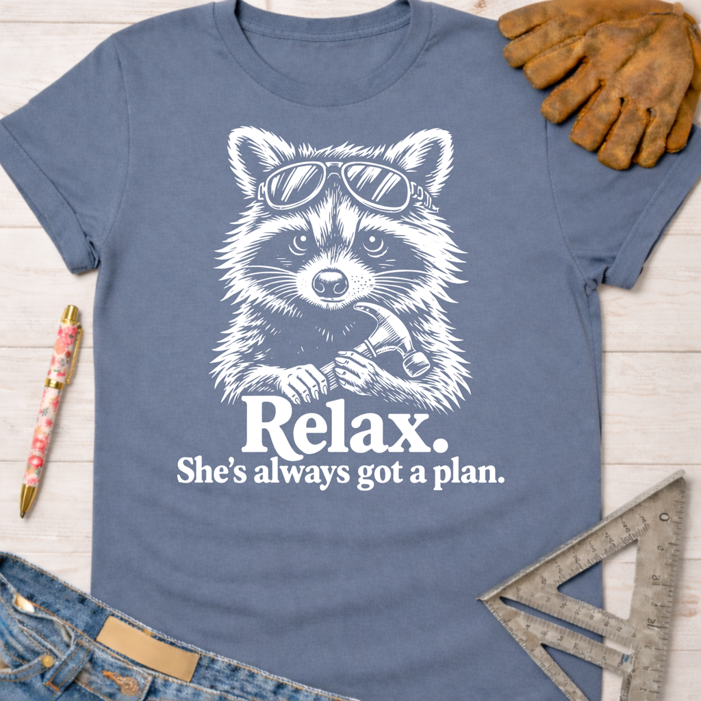 Women's funny woodworking graphic tee shirt for female woodworkers, makers, and DIY builders — Sawdust & Ponytails. Blue cotton tee with a white graphic of a raccoon in sunglasses holding a hammer, text "Relax. She's always got a plan.