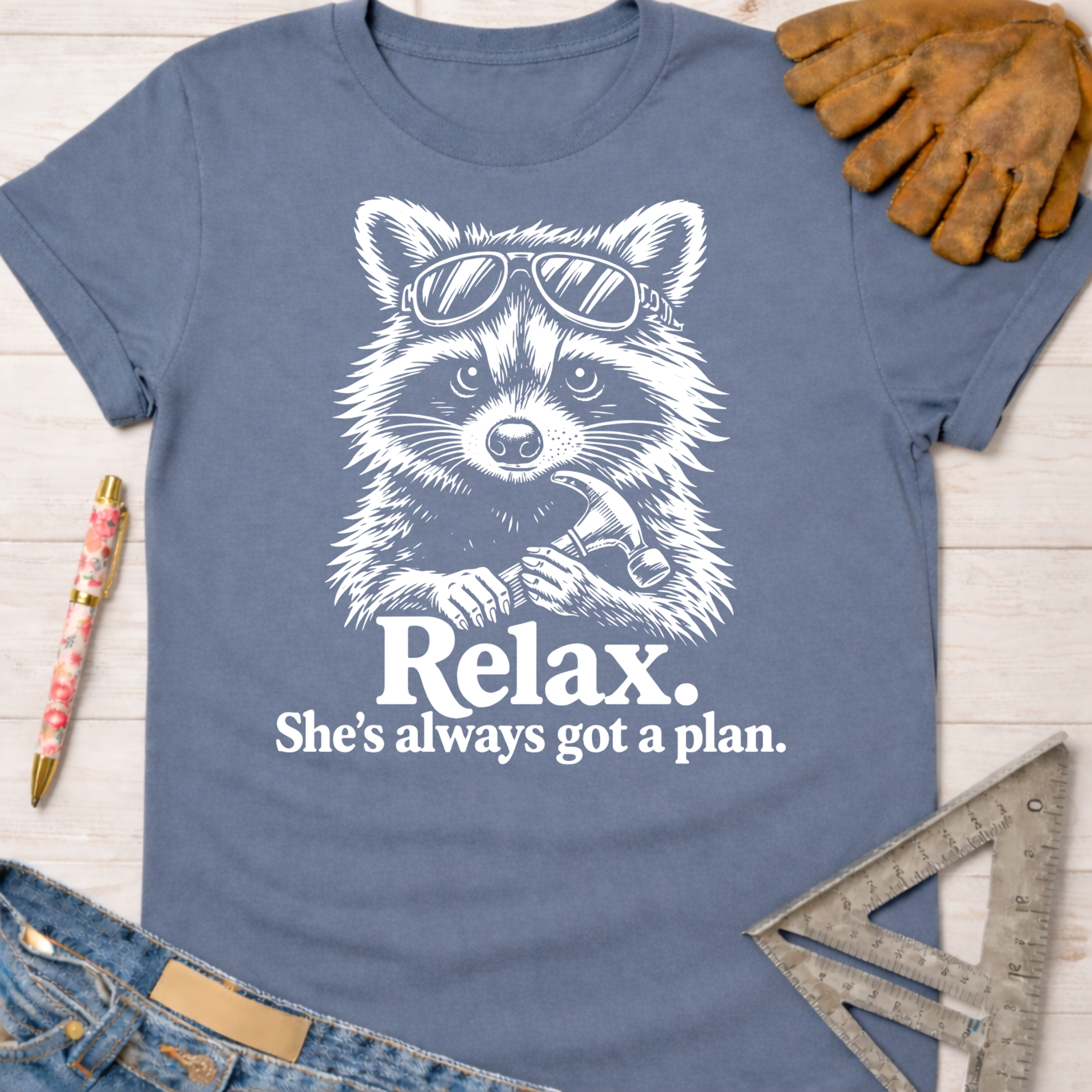 Women's funny woodworking graphic tee shirt for female woodworkers, makers, and DIY builders — Sawdust & Ponytails. Blue cotton tee with a white graphic of a raccoon in sunglasses holding a hammer, text "Relax. She's always got a plan.