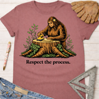 Women's funny woodworking graphic tee shirt for female woodworkers, makers, and DIY builders — Sawdust & Ponytails. Mauve cotton tee with Bigfoot sanding wood on a tree stump, "Respect the process." text.