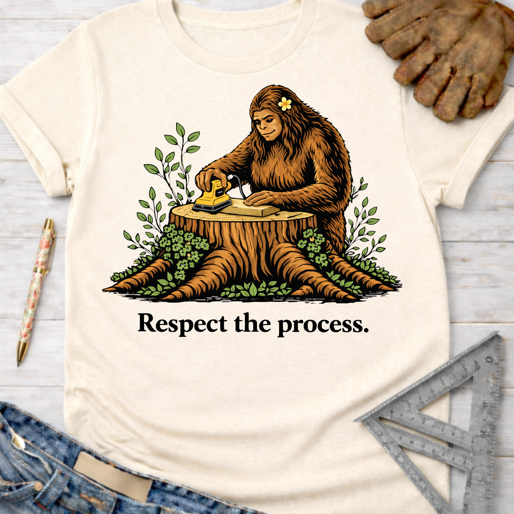 Women's funny woodworking graphic tee shirt for female woodworkers, makers, and DIY builders — Sawdust & Ponytails. Cream t-shirt with Bigfoot sanding wood on a tree stump, surrounded by green leaves, and "Respect the process." text.