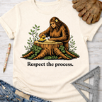 Women's funny woodworking graphic tee shirt for female woodworkers, makers, and DIY builders — Sawdust & Ponytails. Cream t-shirt with Bigfoot sanding wood on a tree stump, surrounded by green leaves, and "Respect the process." text.